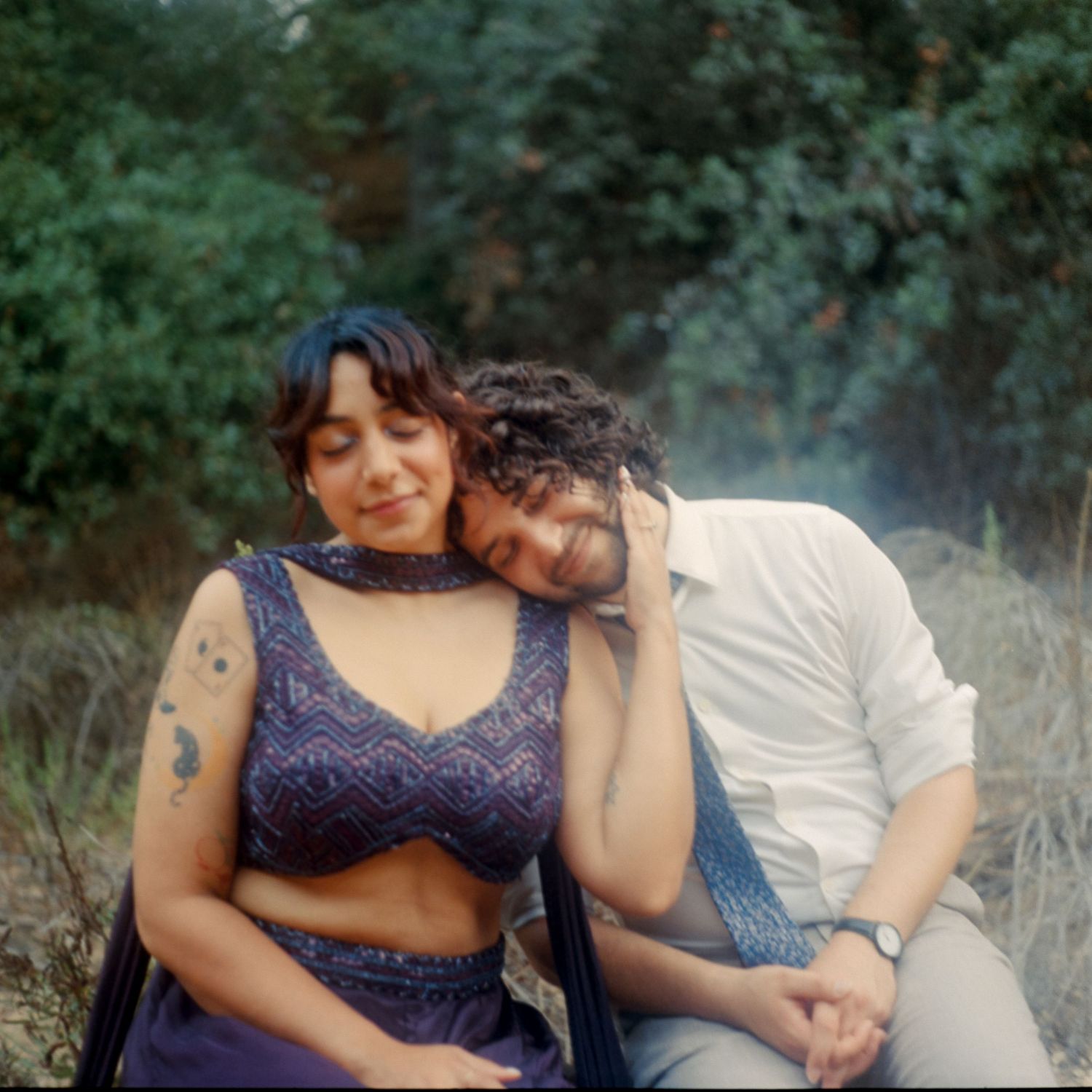 A couple sits together outdoors, woman in blue crop top with tattoo resting head on man's shoulder.