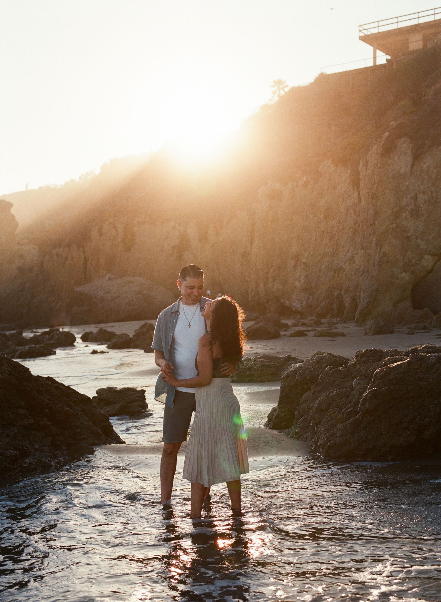 Couple embracing on rocky beach at golden sunset with cliffs and ocean water surrounding them.