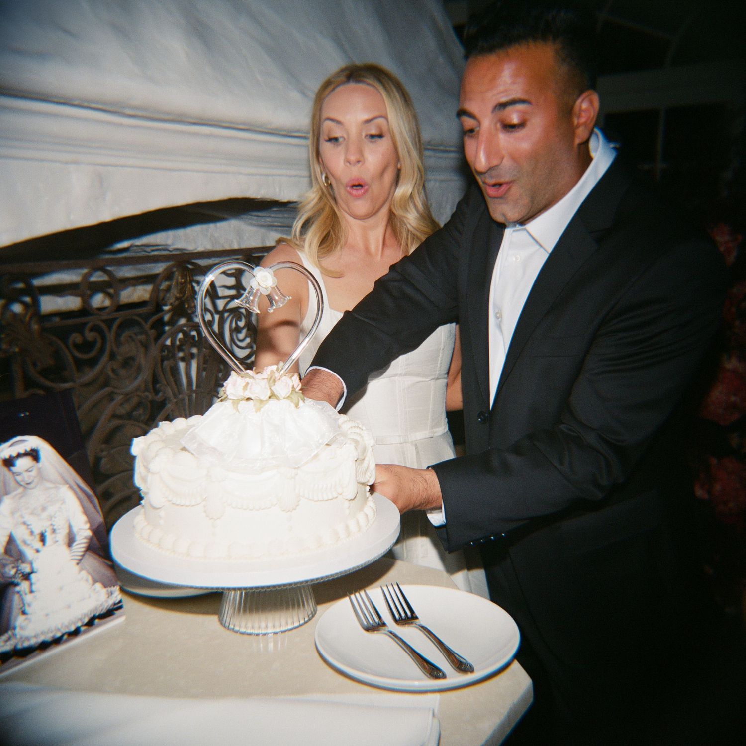 Couple cutting white wedding cake at reception, bride in black dress, groom in suit, forks and plate on table.
