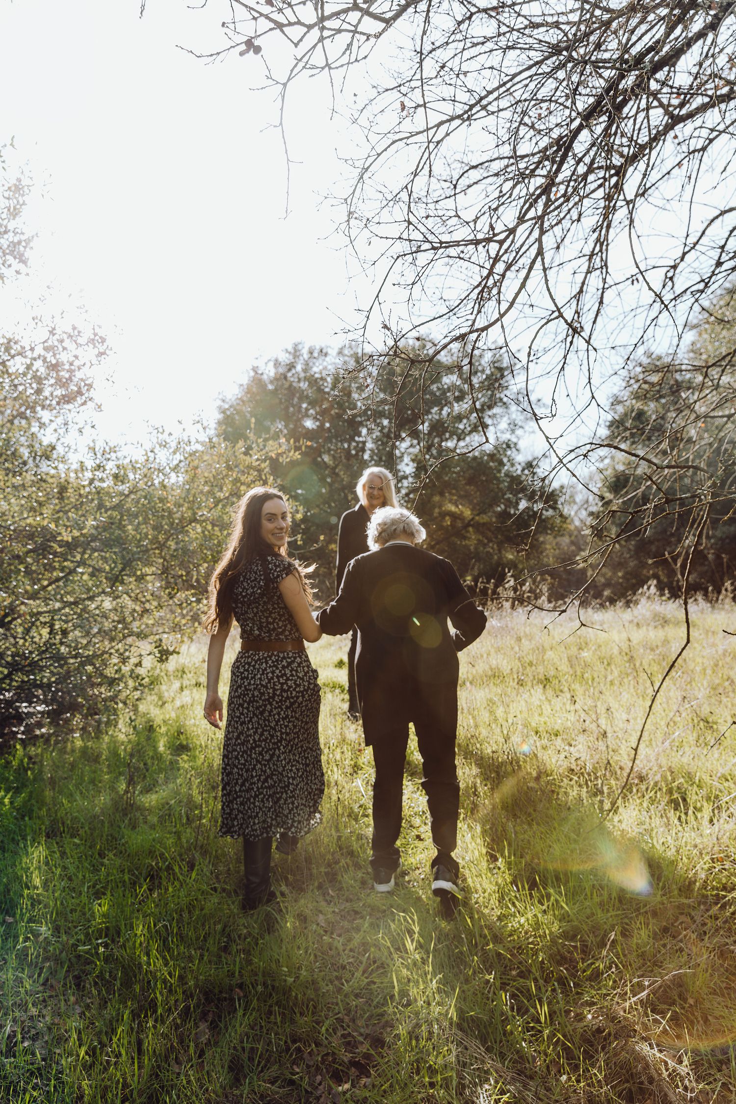 Family walking through sunlit meadow, woman in floral dress holds child as man carries toddler among lush green trees.