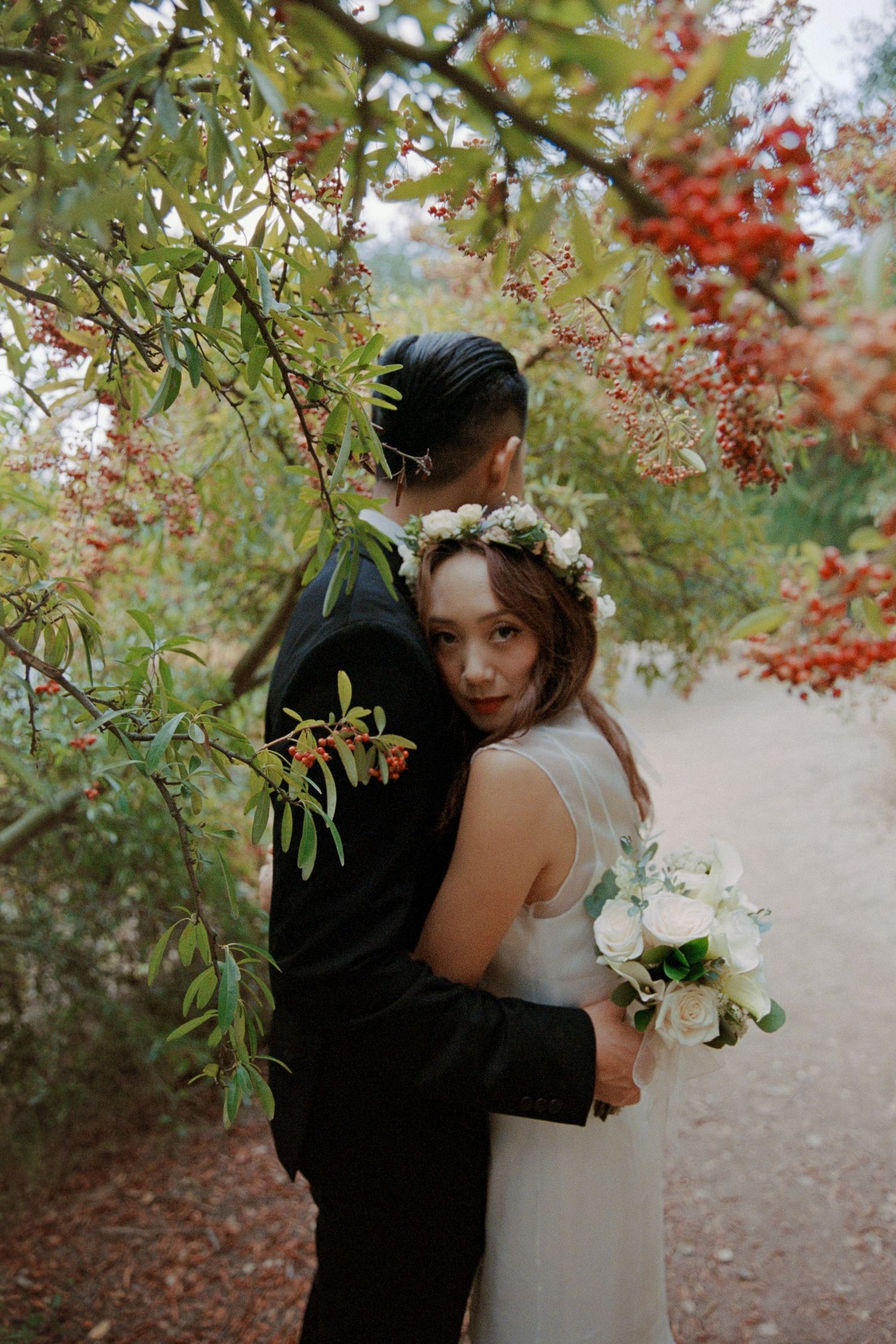 A romantic wedding couple embraces under autumn foliage with red berries and green leaves in a dreamy outdoor setting.