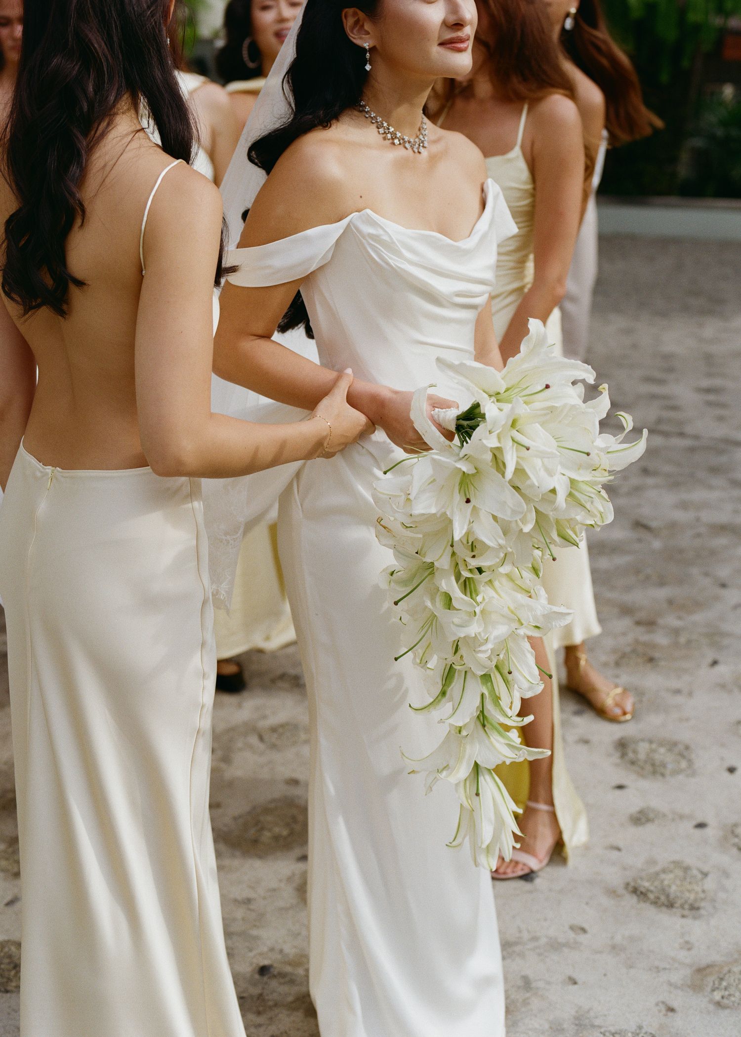 Elegant white cascading lily bouquet held by bride in off-shoulder gown standing with bridesmaids in white dresses.