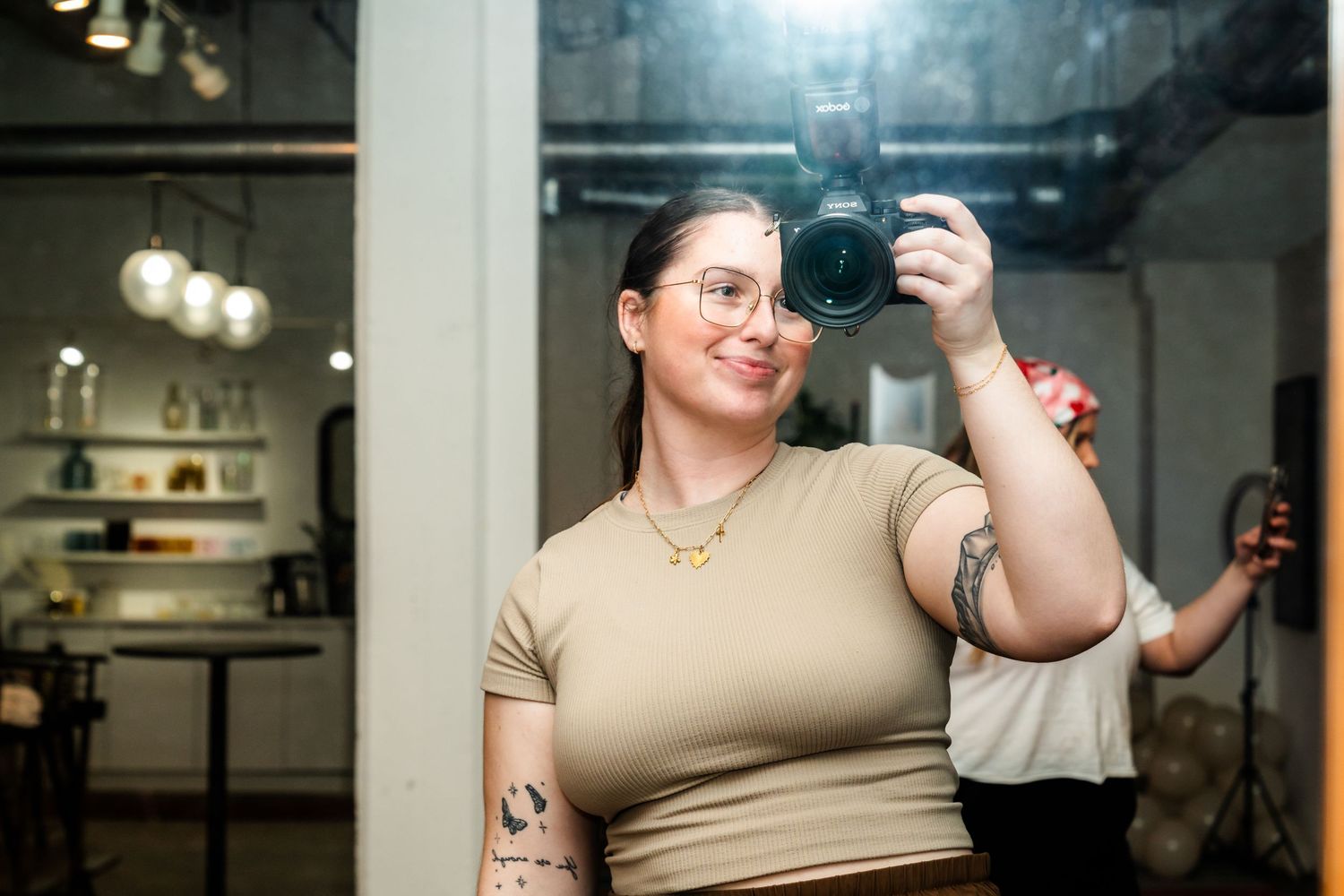 Someone takes a selfie with a professional camera wearing a beige top in an indoor setting with soft lighting.