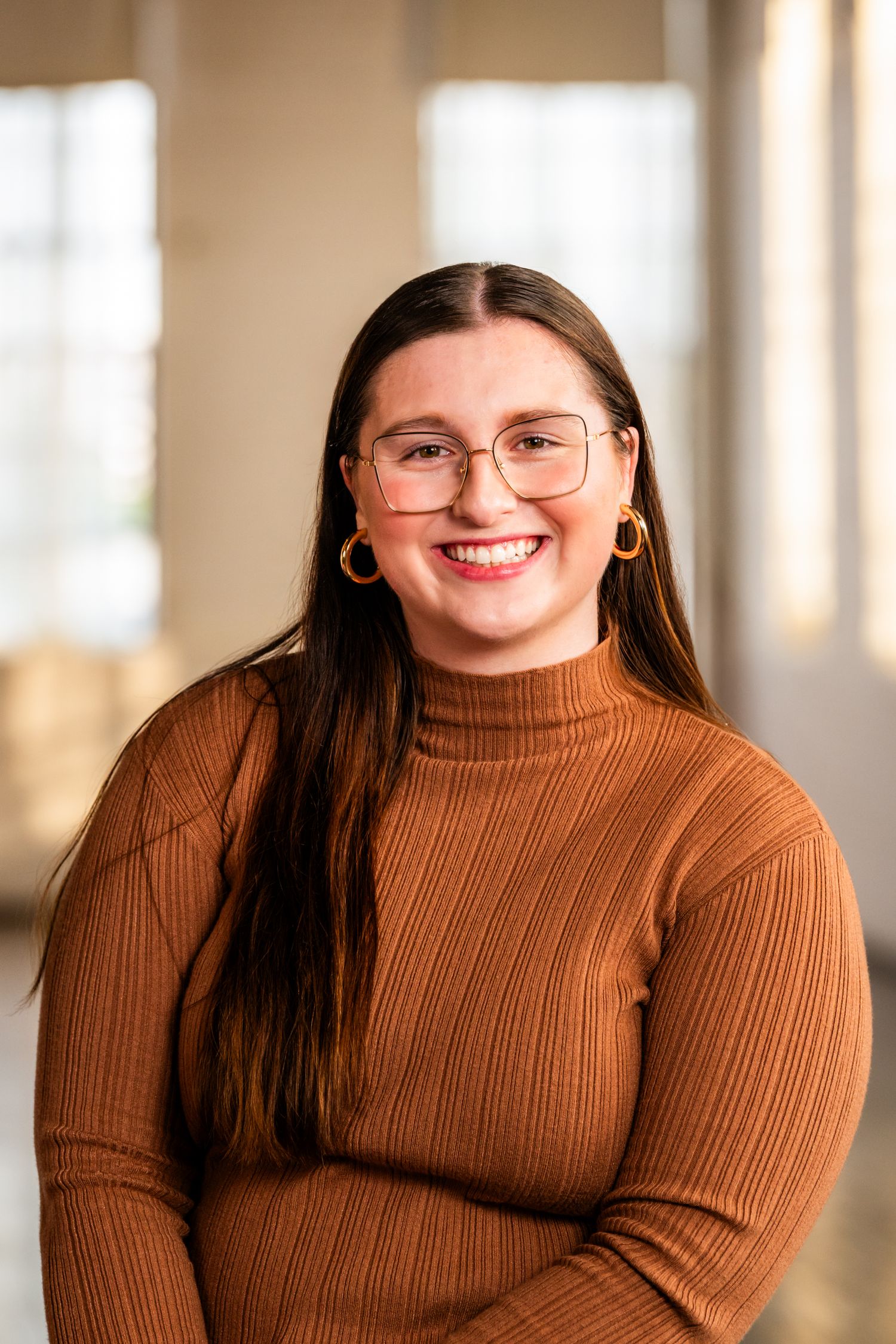 Professional indoor portrait of someone in round glasses and brown turtleneck sweater smiling warmly at camera.