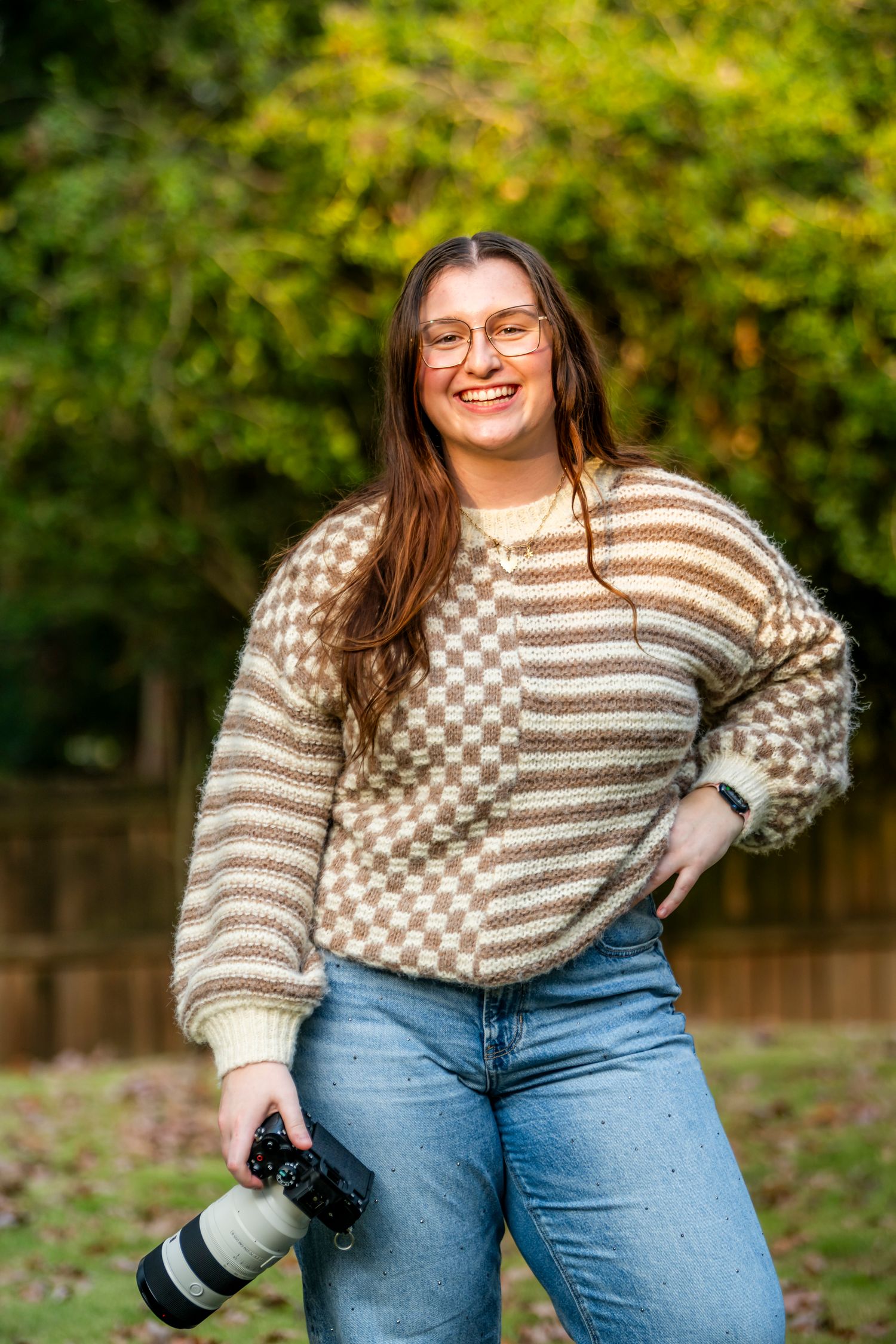 Photographer wearing checkered sweater and jeans holding camera outdoors in casual setting.