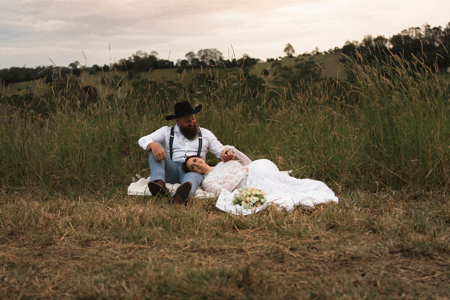 Couple sitting together in a grassy field at sunset, woman in white dress, man in hat and suspenders, picnic basket nearby.