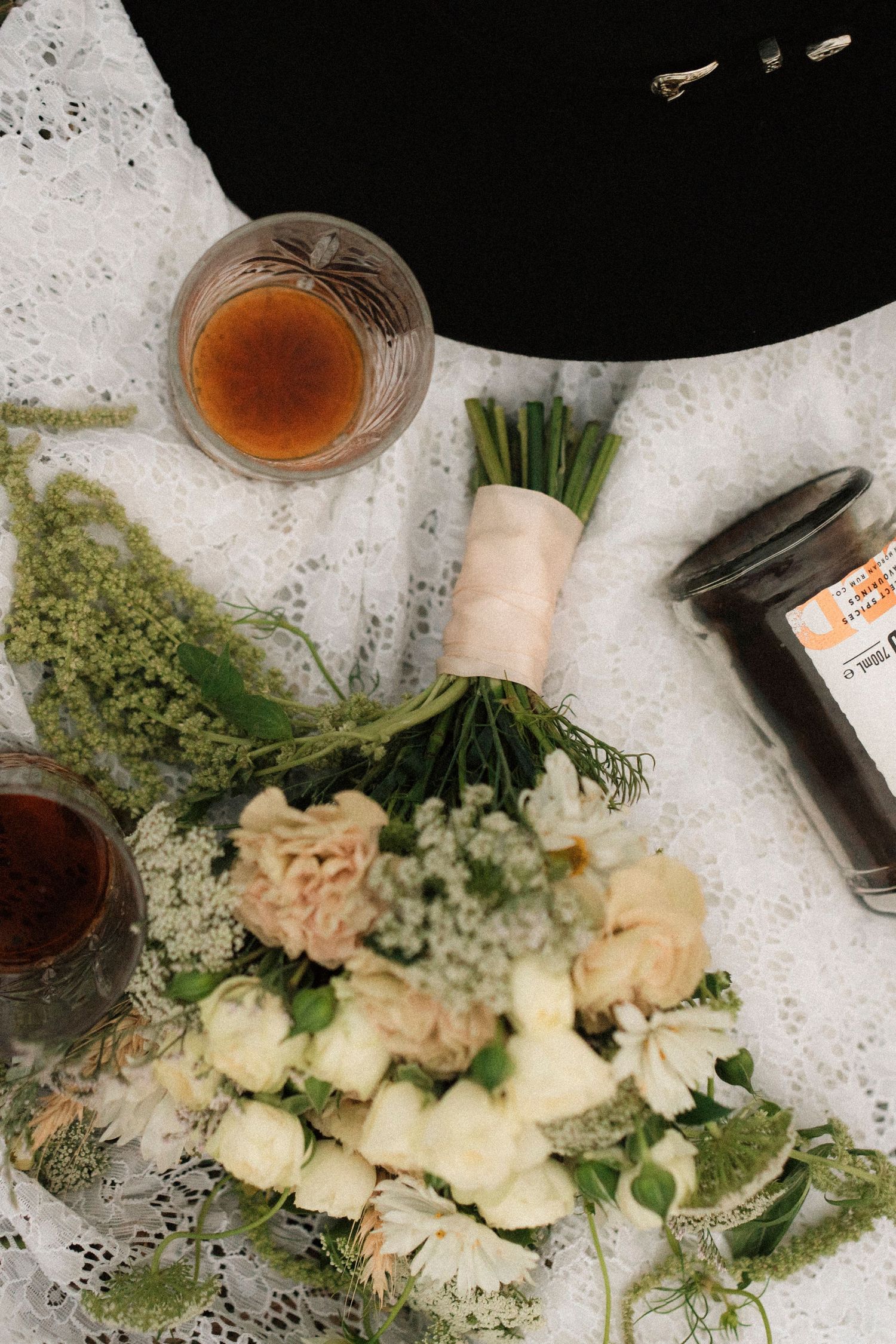 Rustic wedding flat lay with white floral bouquet, cowboy hat, whiskey glasses, and spice jar on lace fabric.