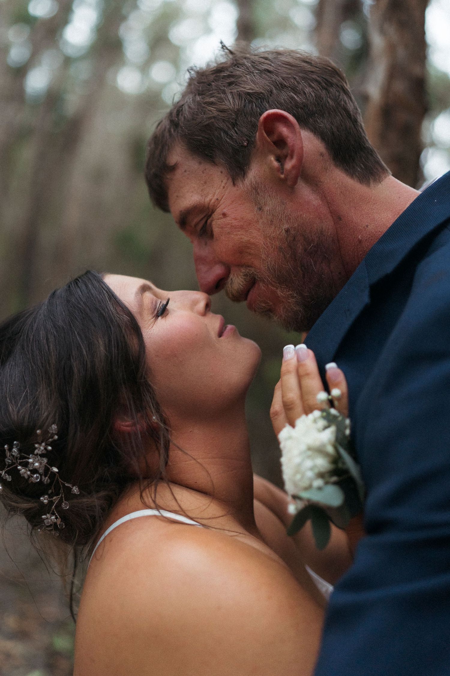 A bride and groom share an intimate moment in a forest setting, with the groom wearing a navy suit and white boutonniere.