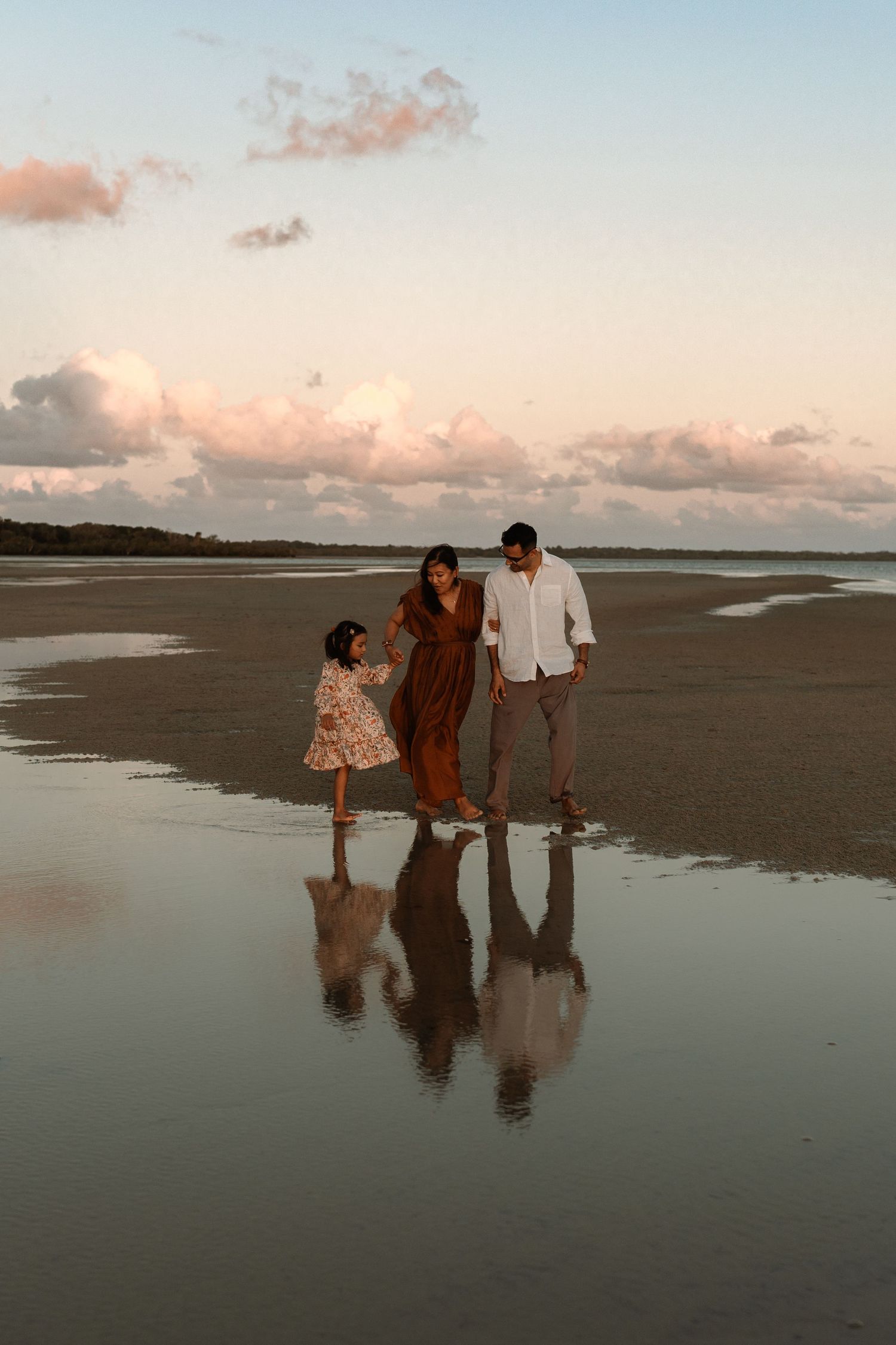 Family of three walking on a reflective beach at sunset, parents holding hands with young daughter.
