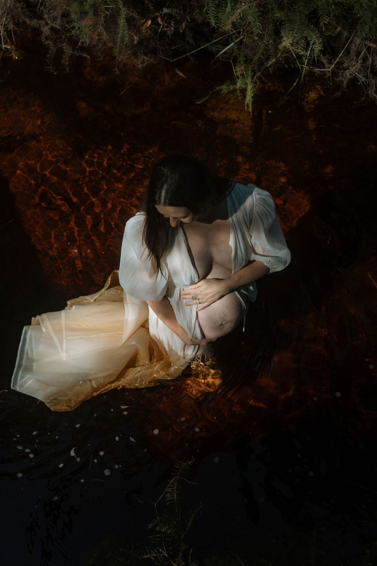 Pregnant woman in sheer white robe crouching near dark water in a moody, atmospheric outdoor maternity photo.