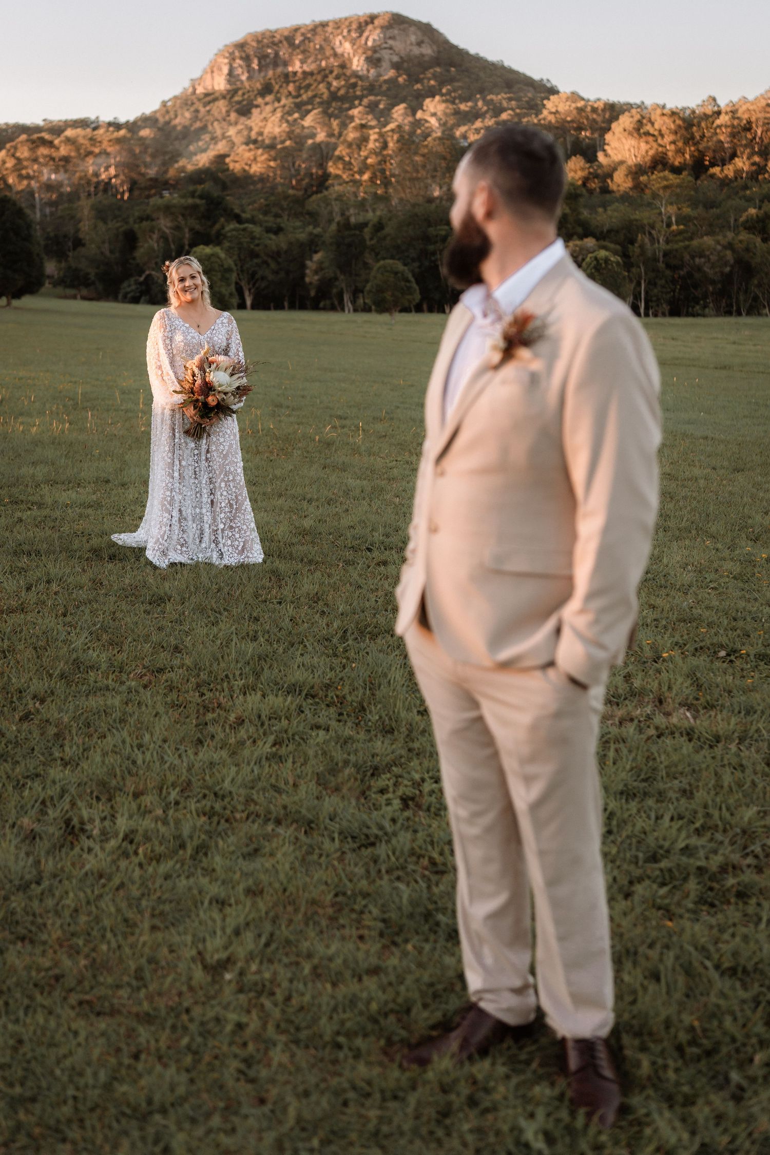 Bride in lace gown holds bouquet as groom in beige suit stands in green field with mountain backdrop.