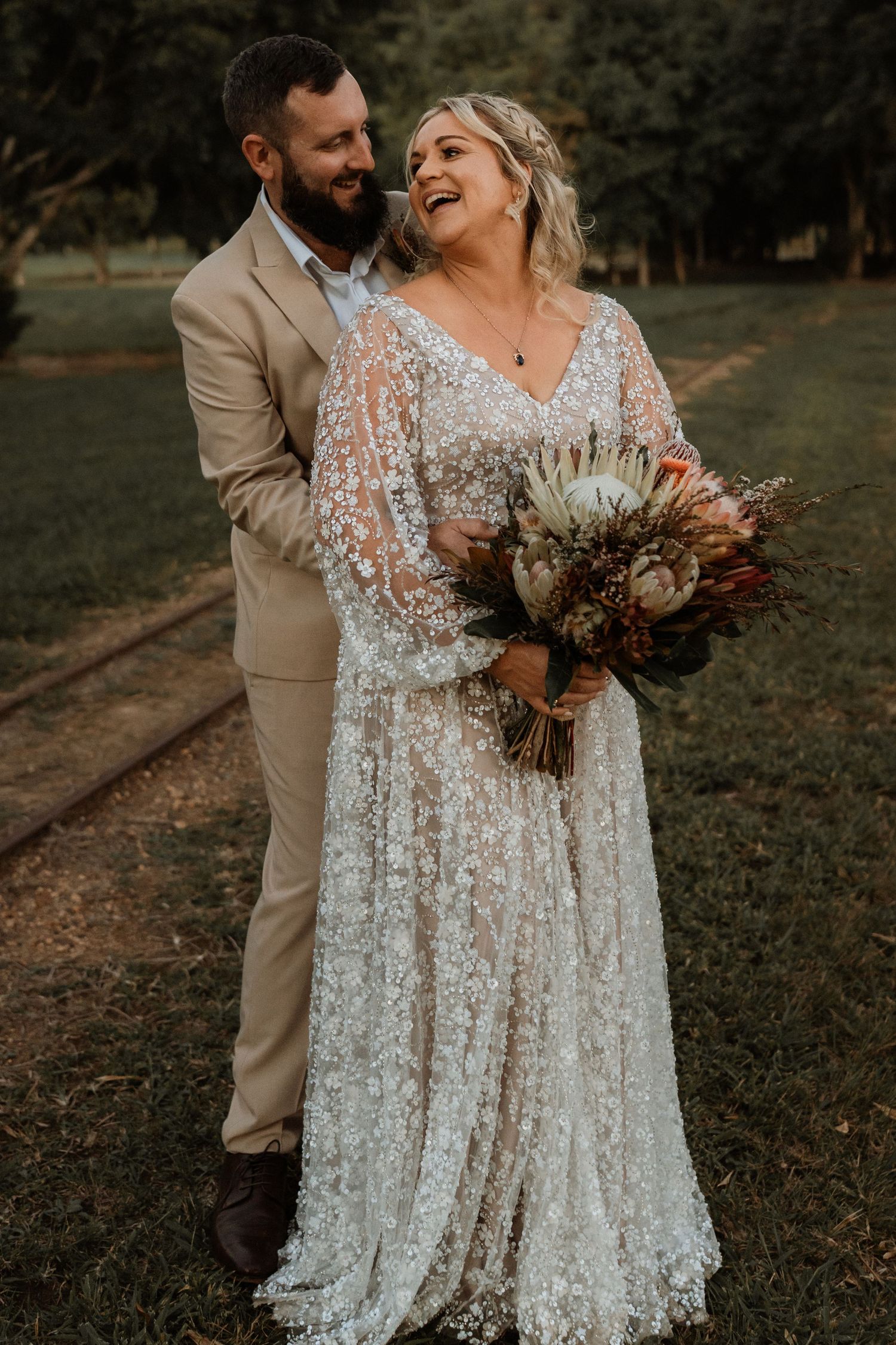Smiling bride in lace gown holding protea bouquet with groom in tan suit outdoors.