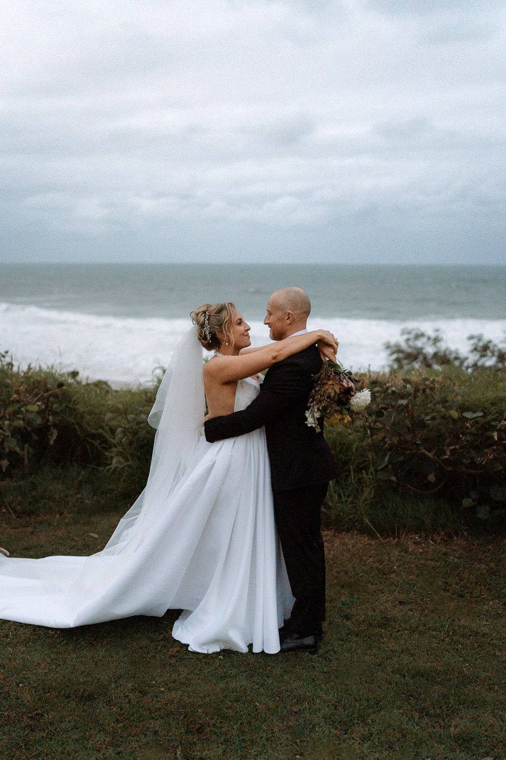 Bride and groom embrace near ocean cliff, she wears white gown with veil, he holds bouquet in black suit.