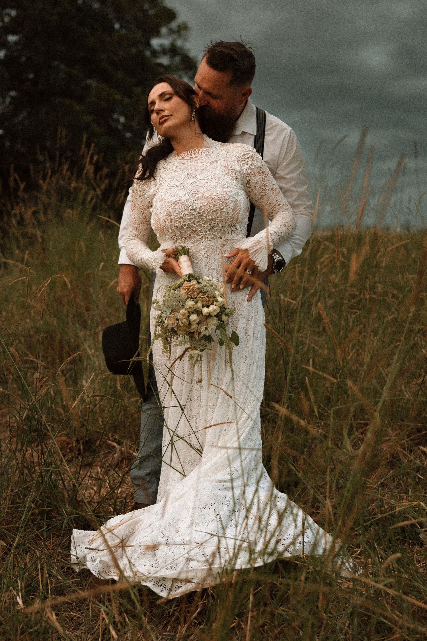Bride in lace gown holds bouquet as groom kisses her cheek in a golden grass field under dramatic cloudy skies.