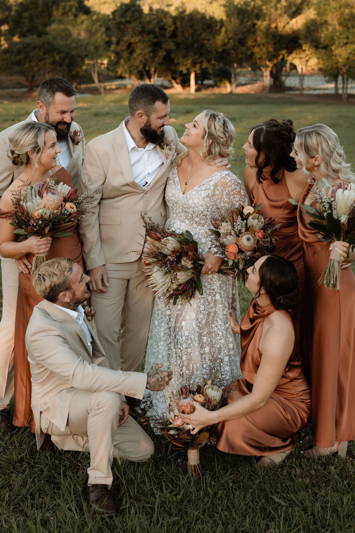 Wedding party in earthy tones, bride in lace gown with groom in tan suit, surrounded by bridesmaids in rust and bronze dresses.
