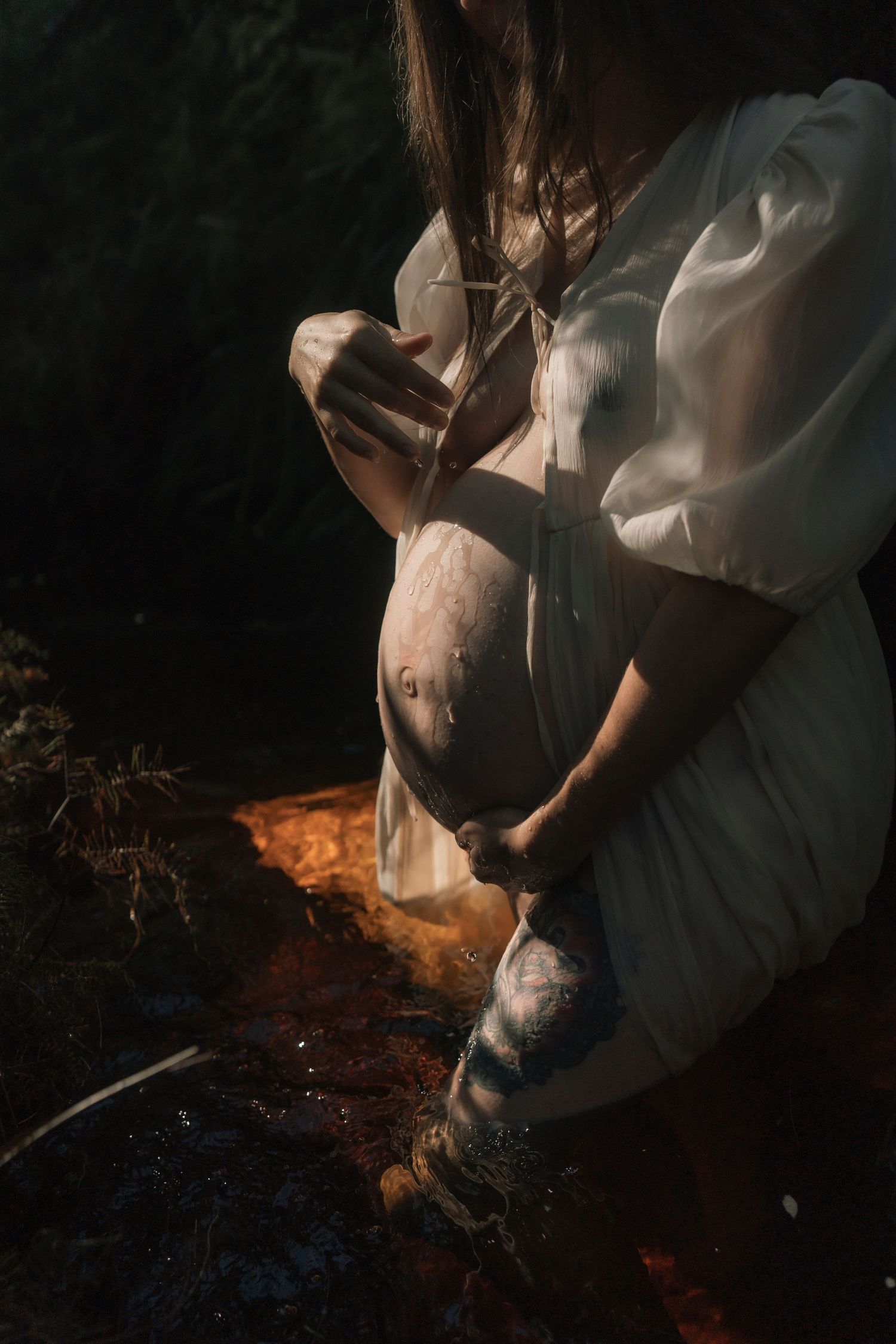 Pregnant woman in white dress cradling baby bump outdoors in moody natural light setting.