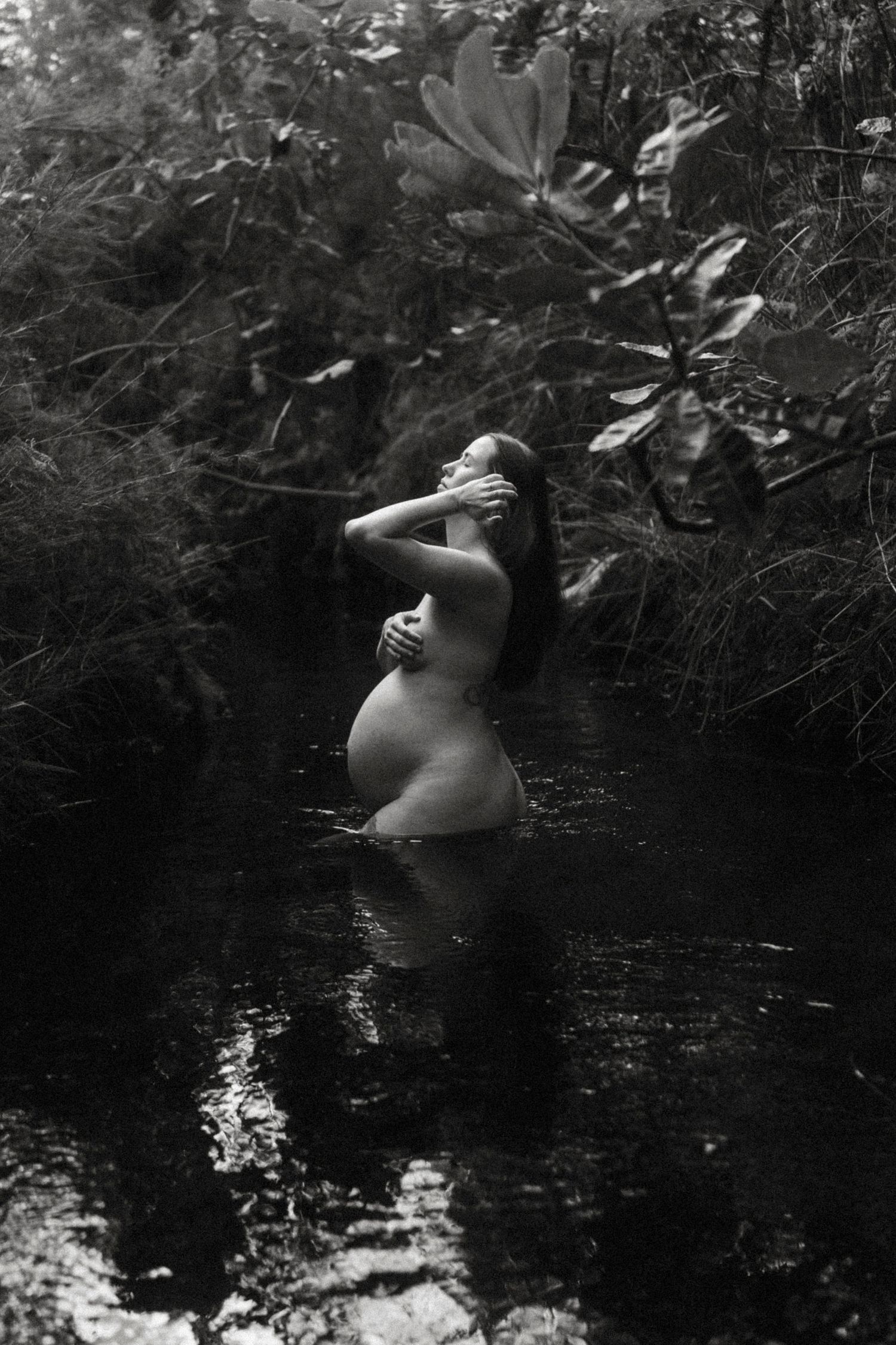 Black and white photo of a pregnant woman standing in a serene forest stream, cradling her belly.