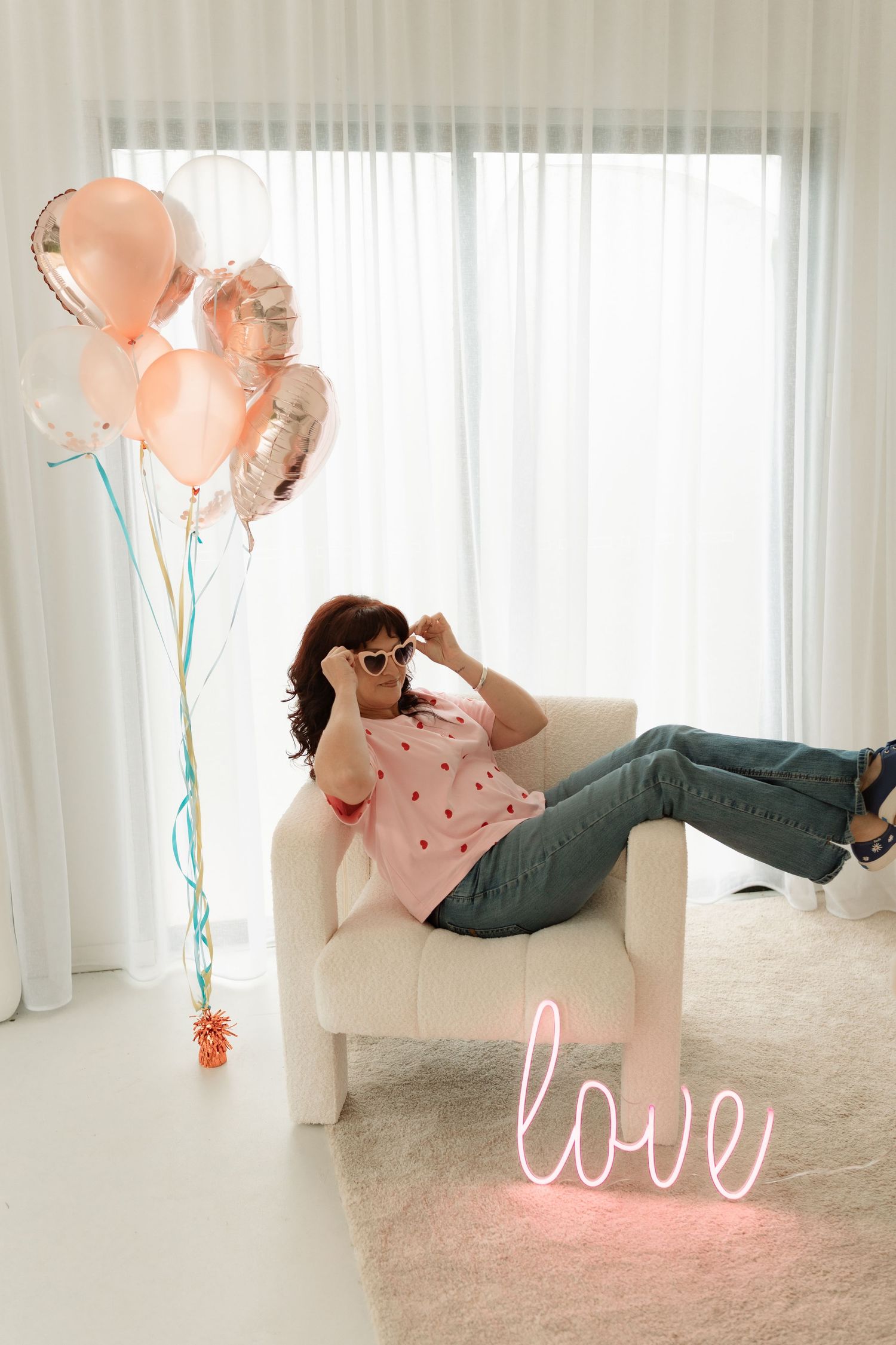 Woman relaxing in white armchair with pink balloons and neon love sign in bright modern room.