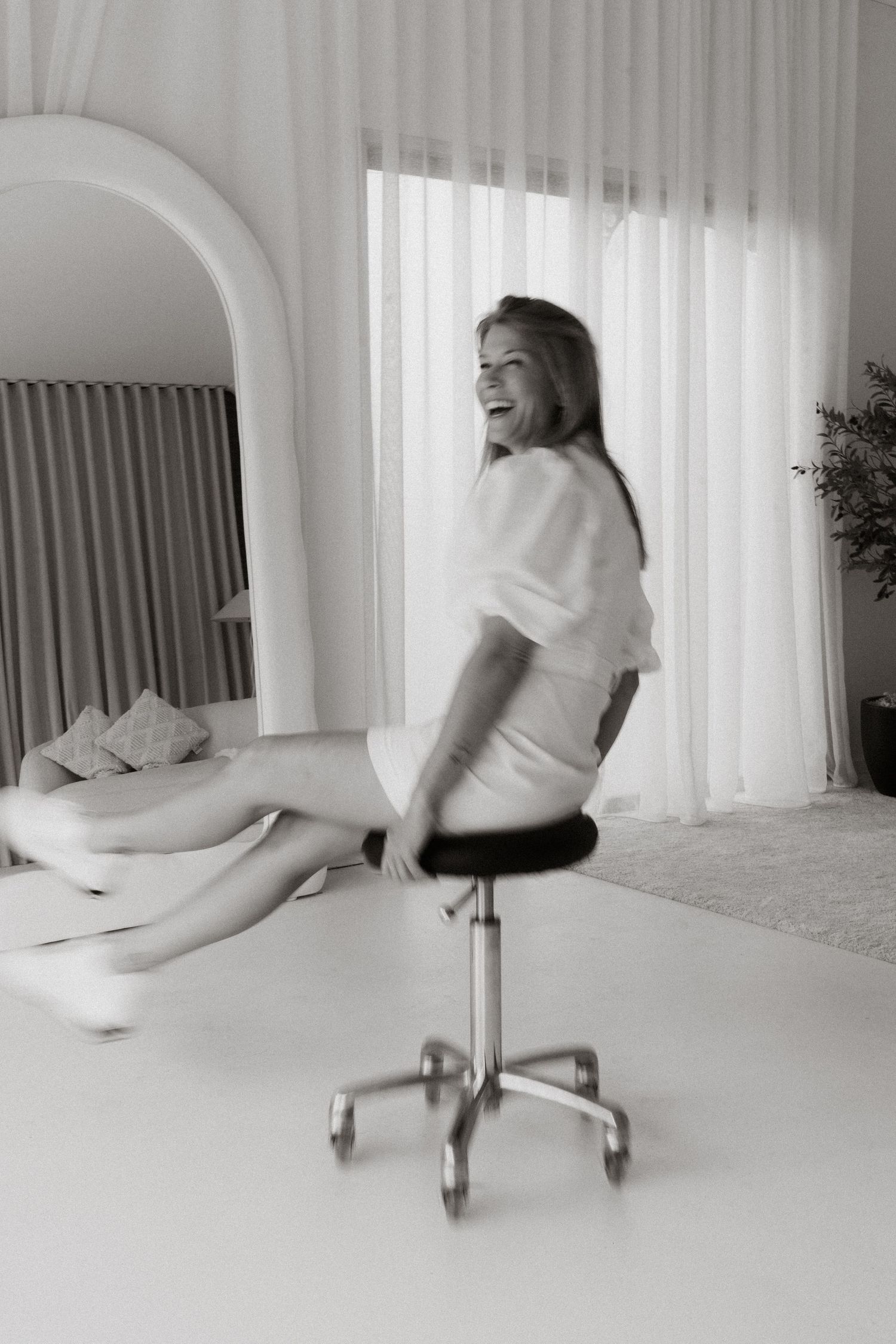 Black and white photo of a laughing woman spinning on a office chair in a bright, modern living room.