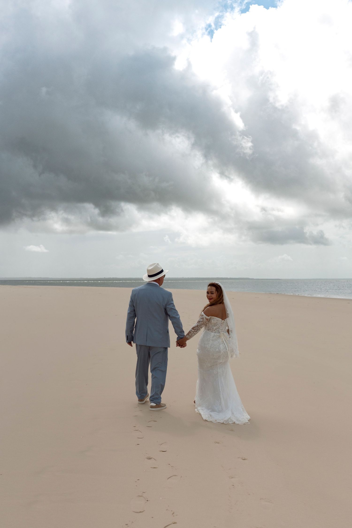 Bride and groom holding hands walking on sandy beach with dramatic cloudy sky, wedding couple in white dress and gray suit.