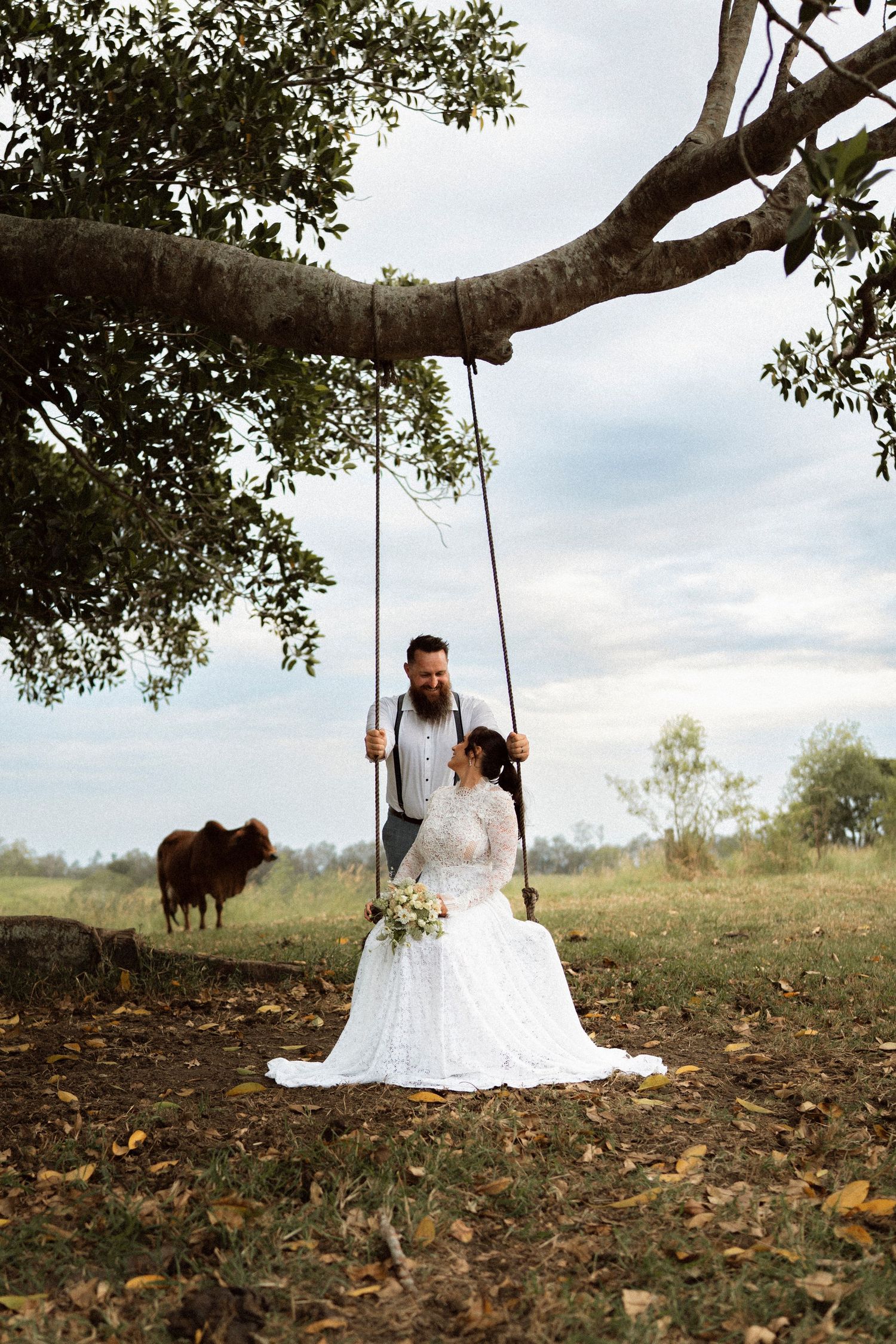 Bride in lace gown sits on tree swing as groom stands behind her, with a cow grazing in a rustic field backdrop.