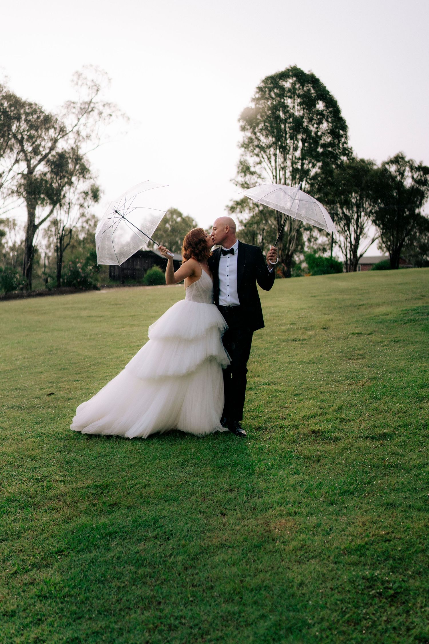 A couple in formal wedding attire shares a romantic dance together on a grassy hillside at sunset.