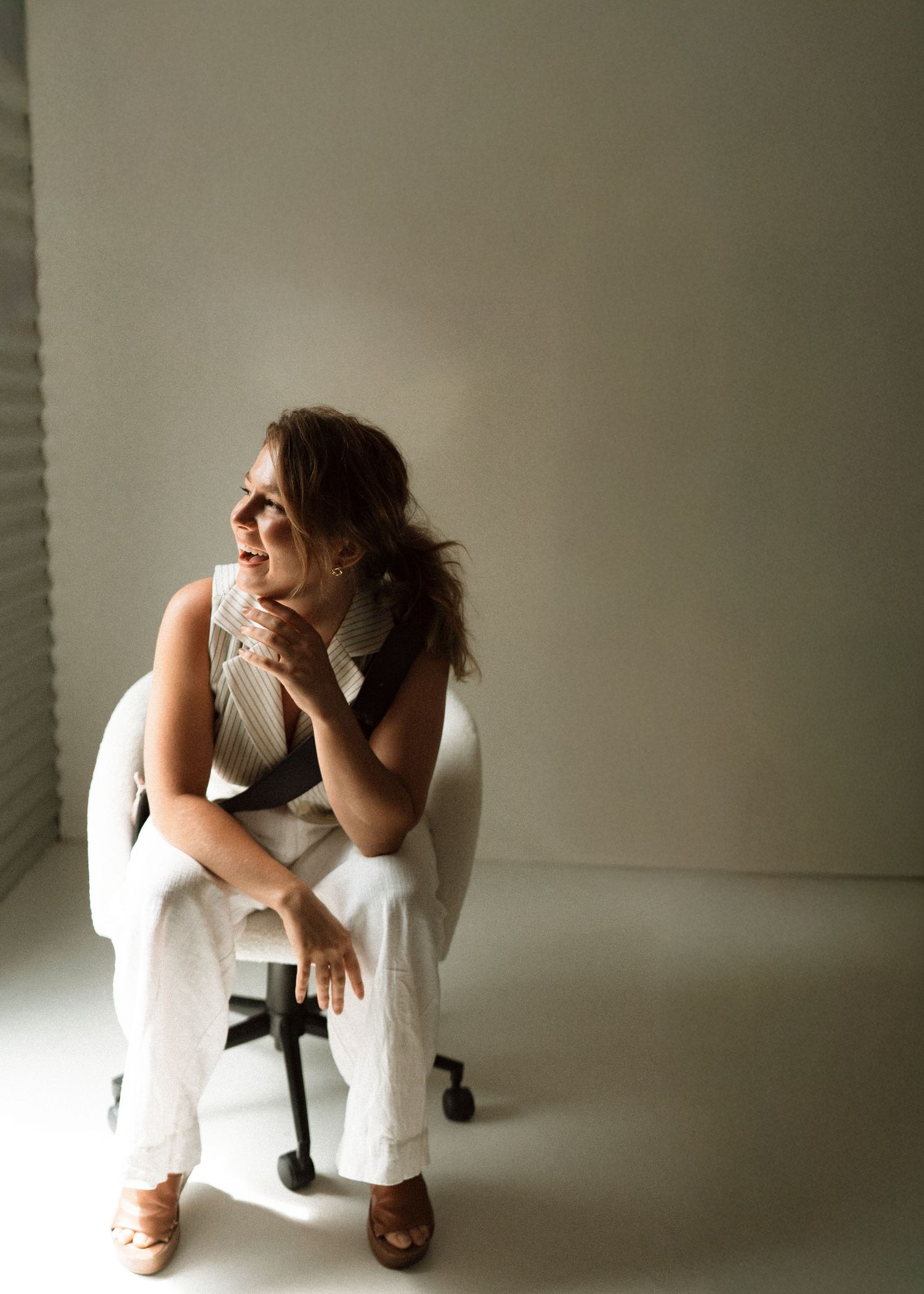 Woman in white outfit sitting on modern chair, smiling and looking away in bright minimalist studio setting.