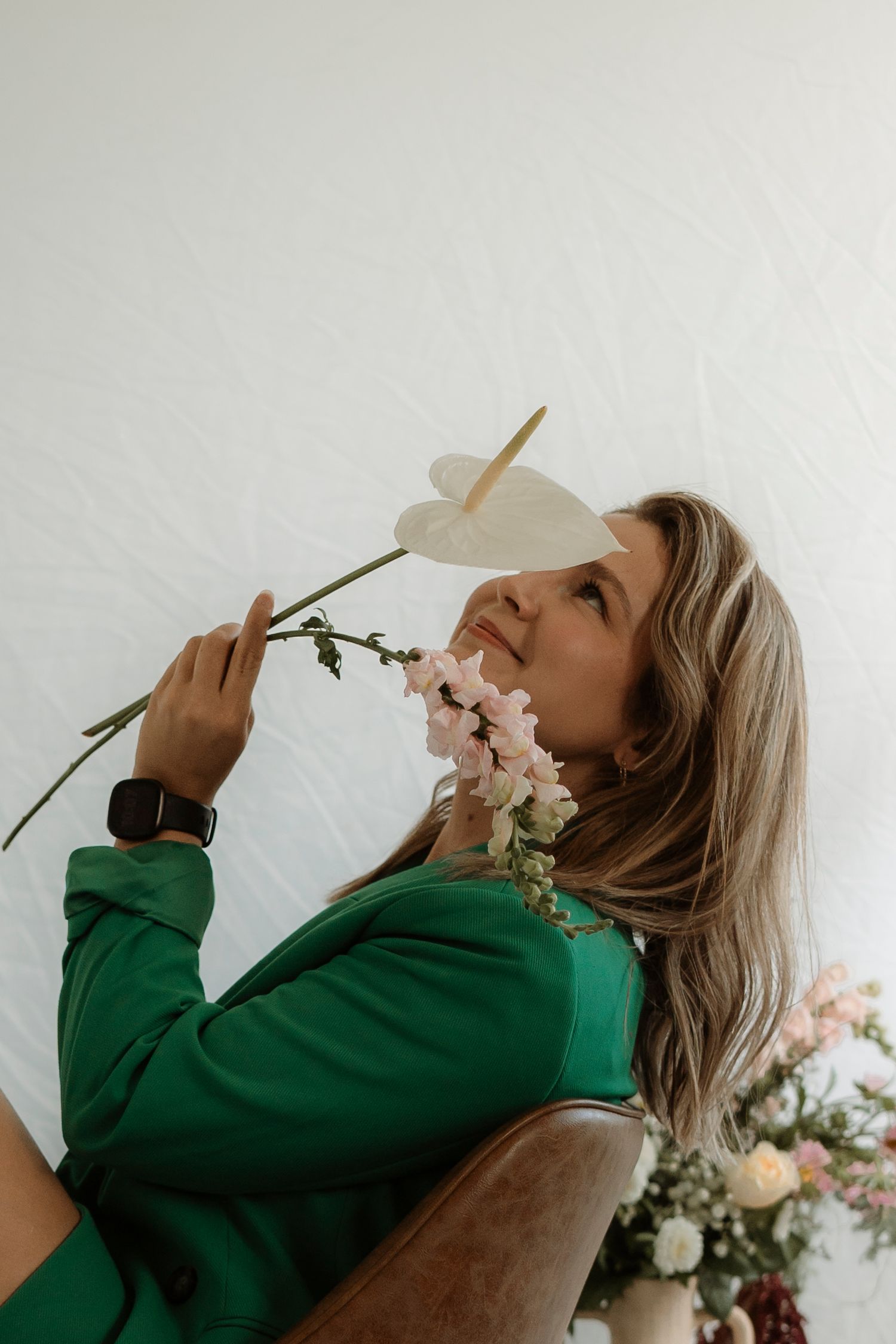 A person in a green sweater sits in a brown chair holding delicate pink flowers against a white wall. personal branding session