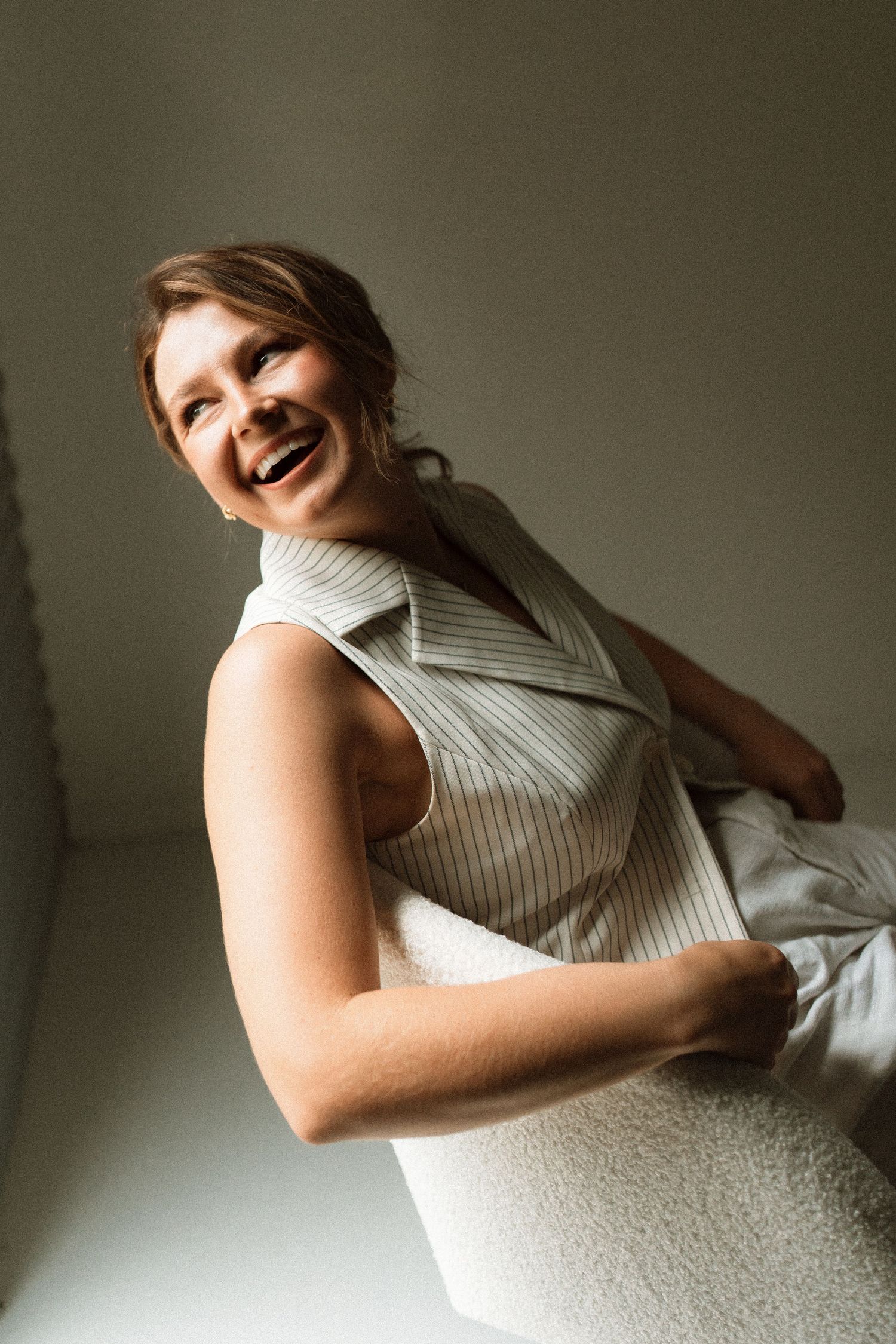 A cheerful woman in a striped sleeveless top laughs joyfully while leaning against a white surface indoors.