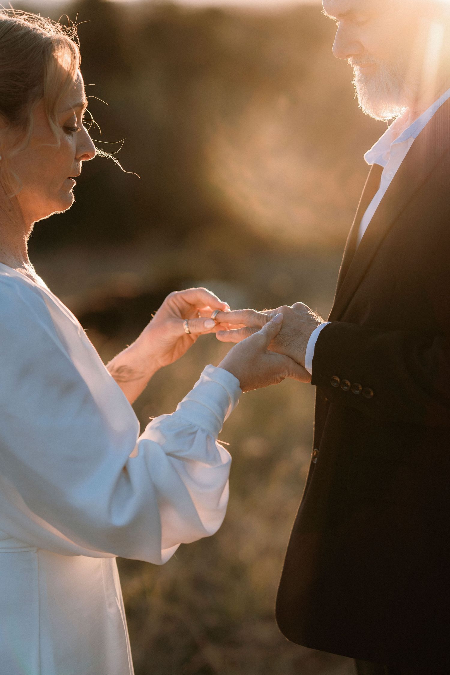 Bride placing wedding ring on groom's finger during golden hour outdoor ceremony, backlit by warm sunset light.