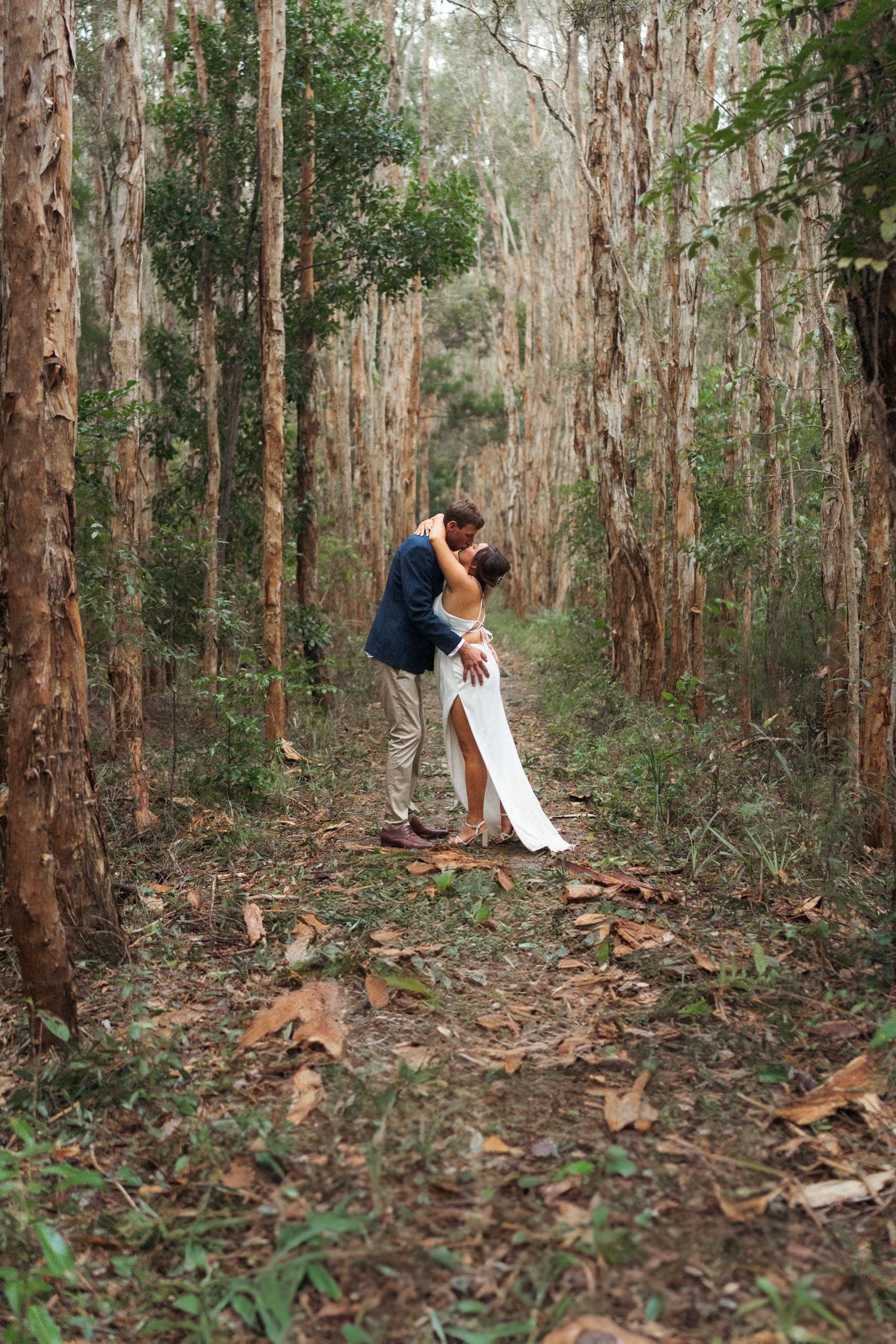 A couple shares a romantic kiss in a lush forest setting, the bride in a white gown and groom in a navy suit.