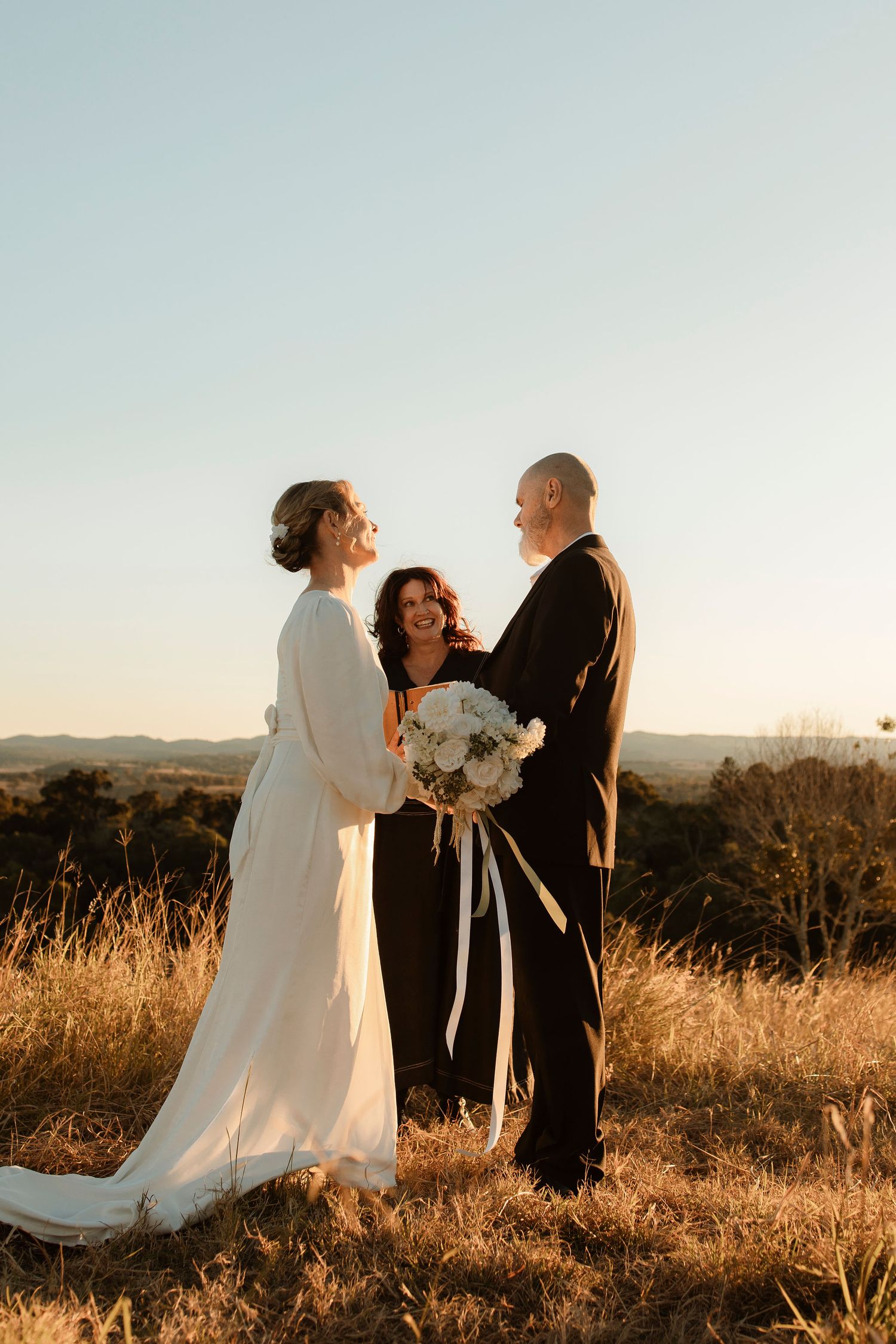 Bride in white gown and groom in black suit exchange vows with officiant at golden hour outdoor wedding ceremony.