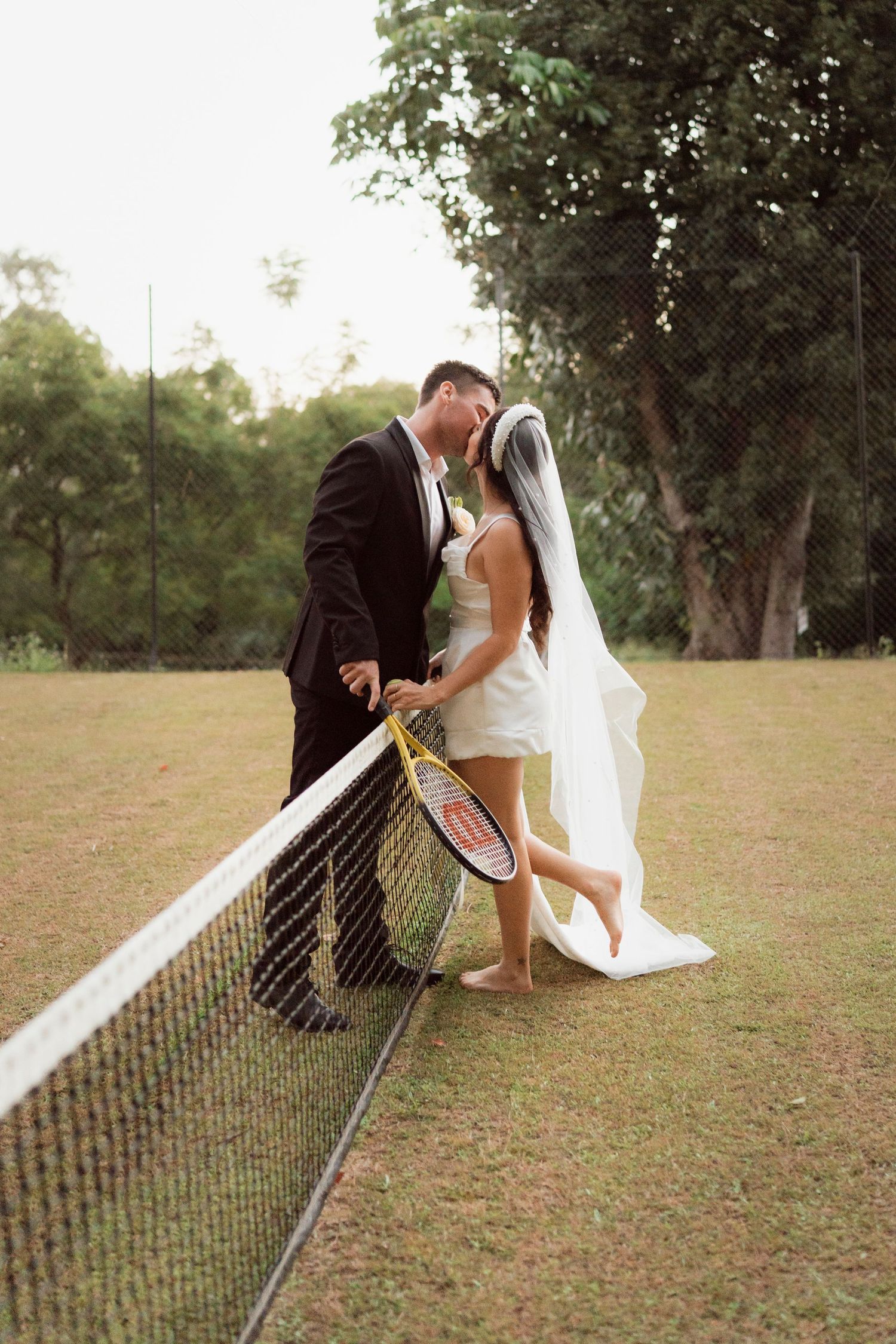 Bride and groom kissing at tennis net, she holds a racket wearing a short white dress with veil, he wears a black suit.