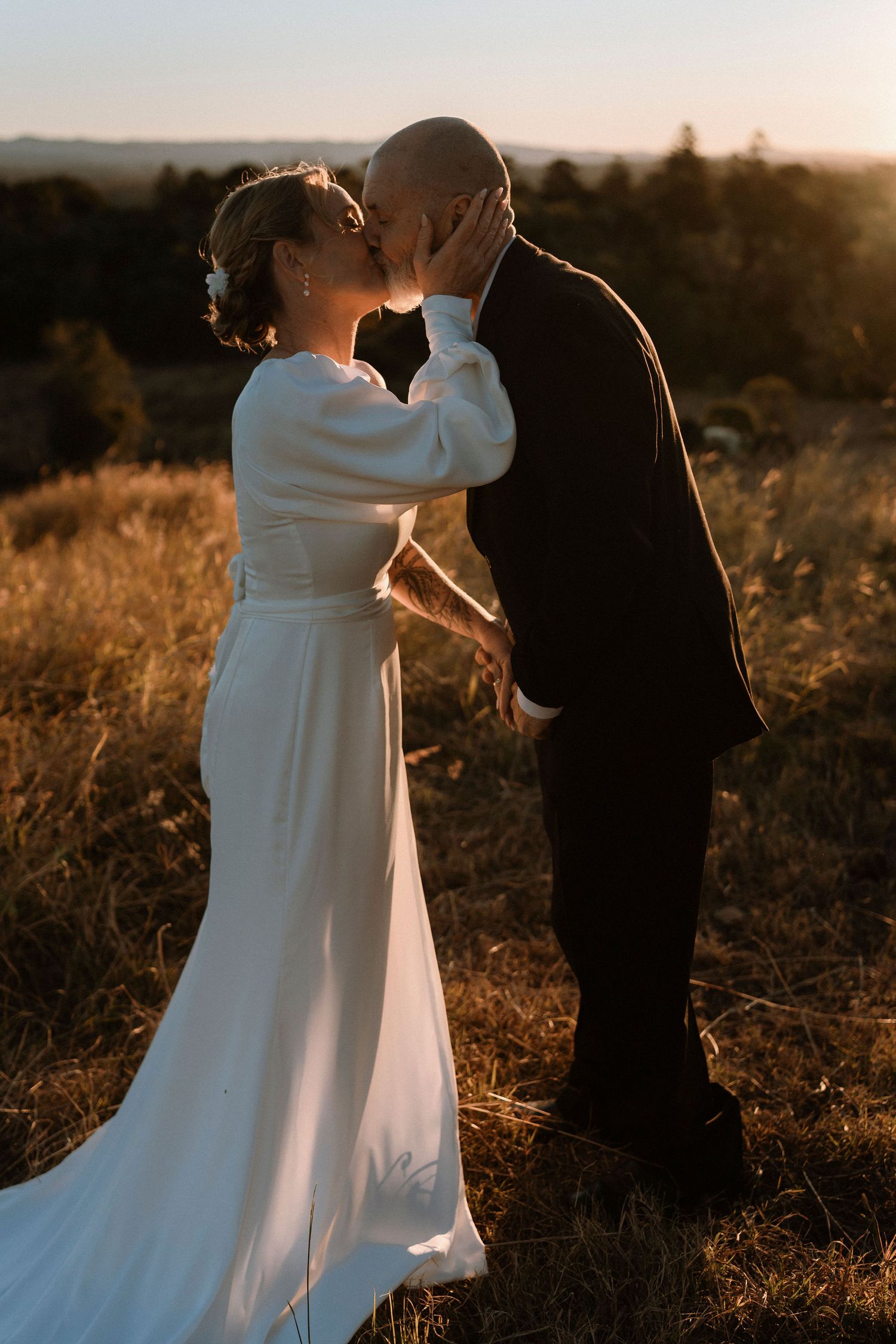 Bride and groom share a romantic sunset kiss in a golden field, bride in elegant white long-sleeve gown.