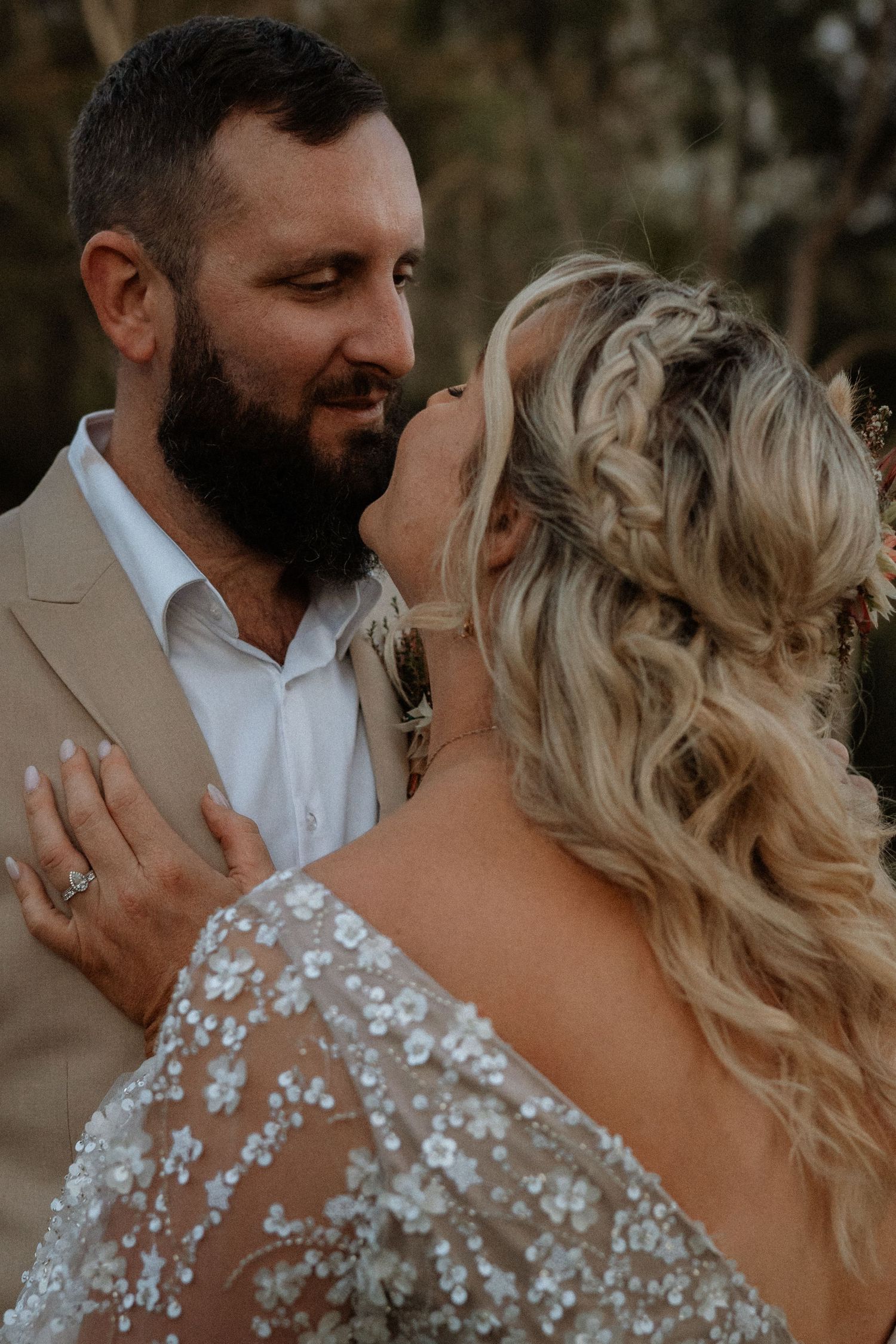 Bride and groom share an intimate moment, bride in lace gown with braided blonde hair, groom in tan suit with beard.