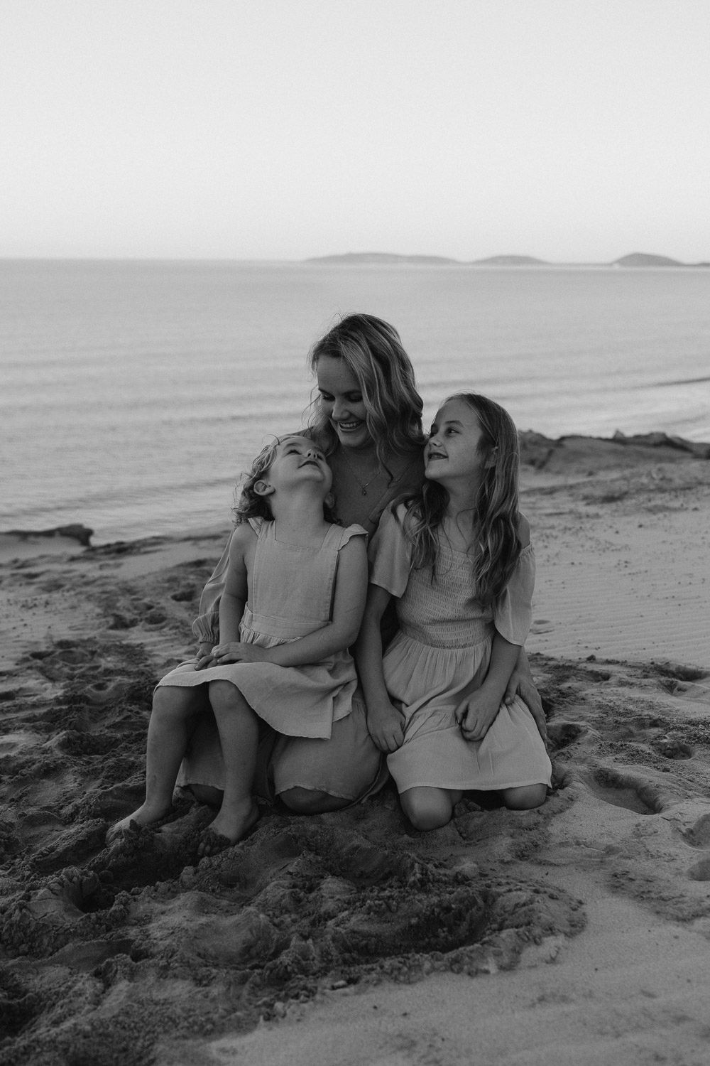 Three figures share a joyful moment together sitting on a beach at sunset in black and white.