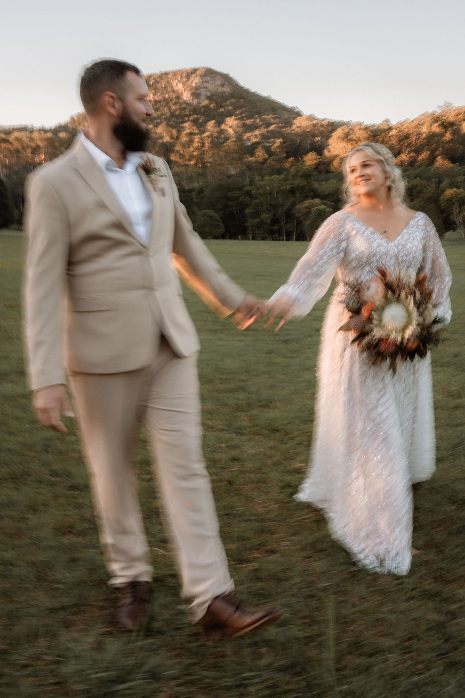Bride and groom hold hands in a sunlit field with mountain backdrop, bride in lace gown holding a boho bouquet.