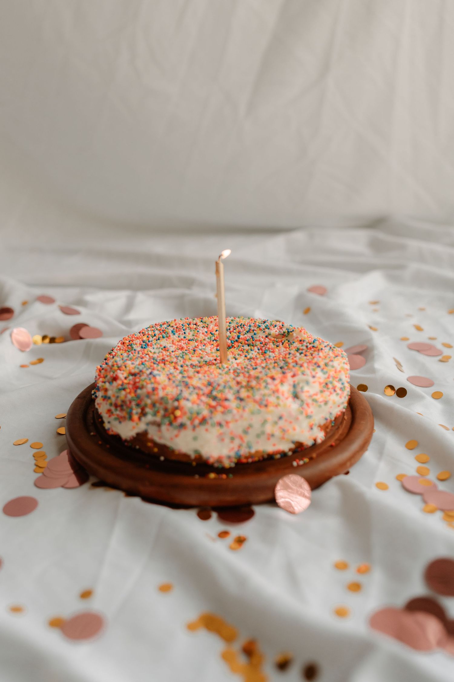 A birthday cake with colorful sprinkles and a lit candle on a wooden stand, surrounded by confetti on white fabric.