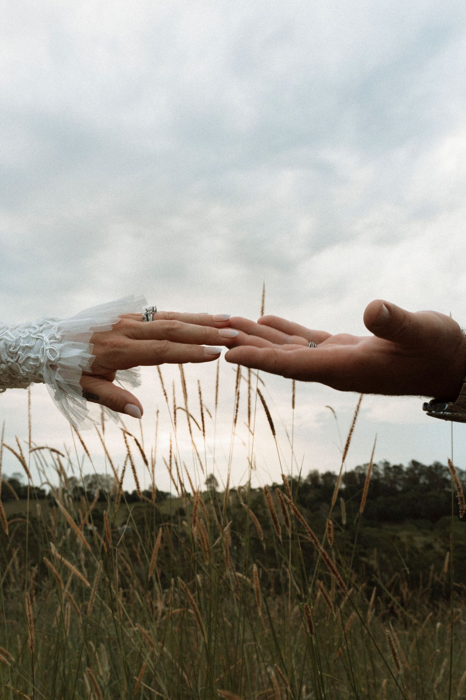 Two hands reaching toward each other in a grassy field, one wearing a wedding ring and lace sleeve, under a cloudy sky.