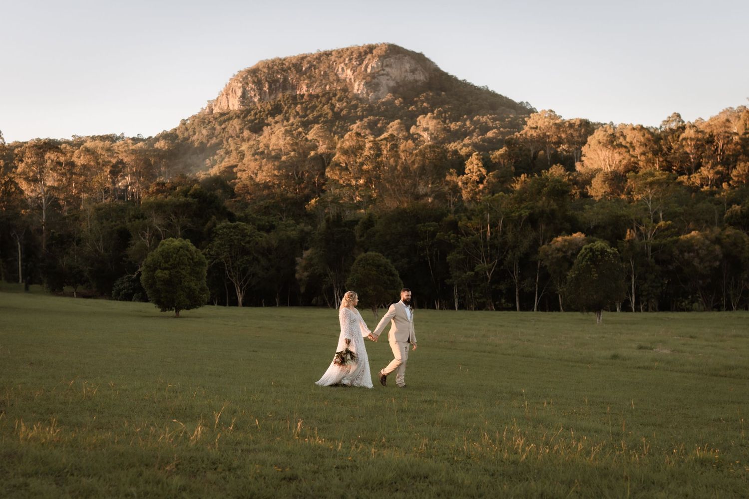 A bride and groom walk hand-in-hand across a sunlit green field with a mountain and trees in the background.