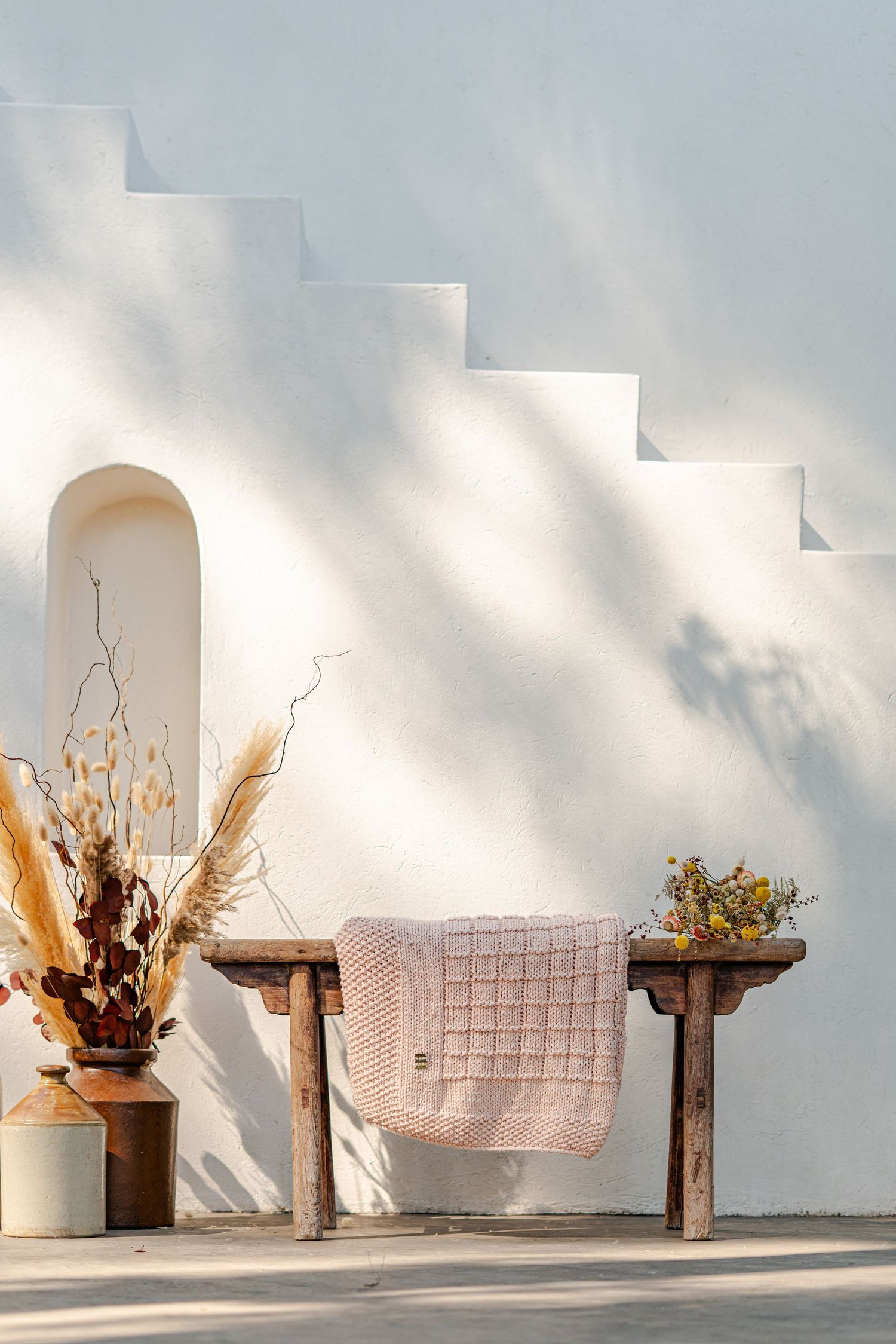 Rustic wooden bench with pink knit blanket against white stucco wall with arched niche and stepped architecture.
