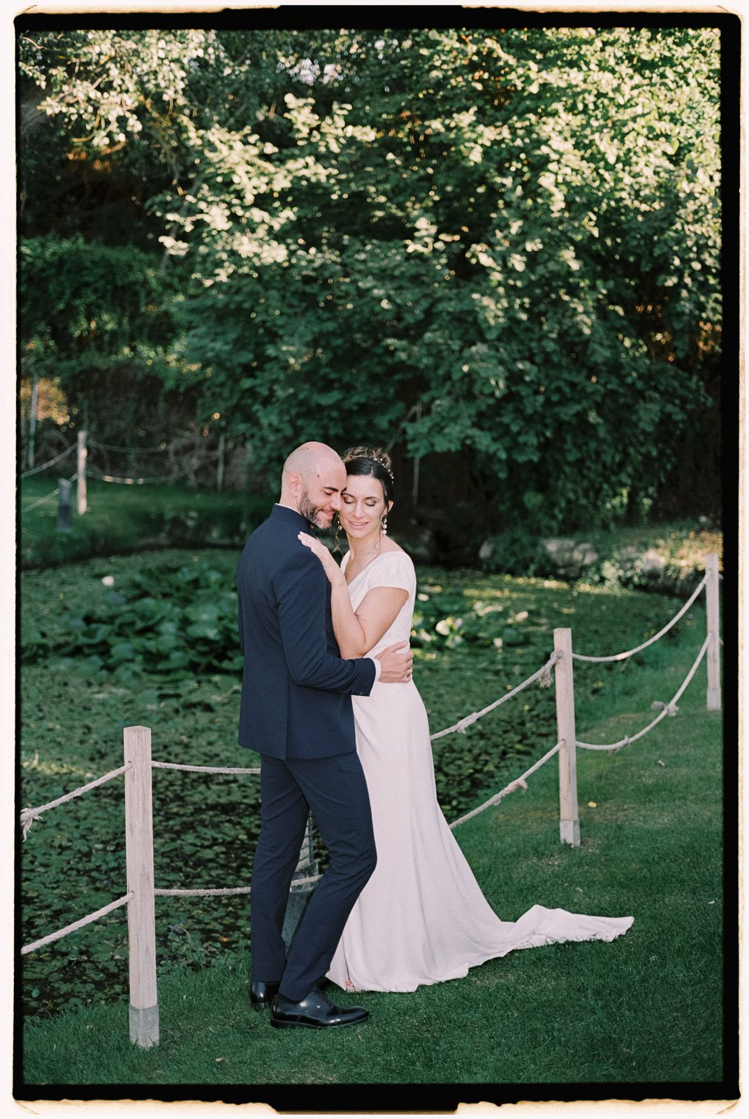 A bride and groom share a tender embrace in a lush green garden, with the bride in a white gown and groom in a dark suit.