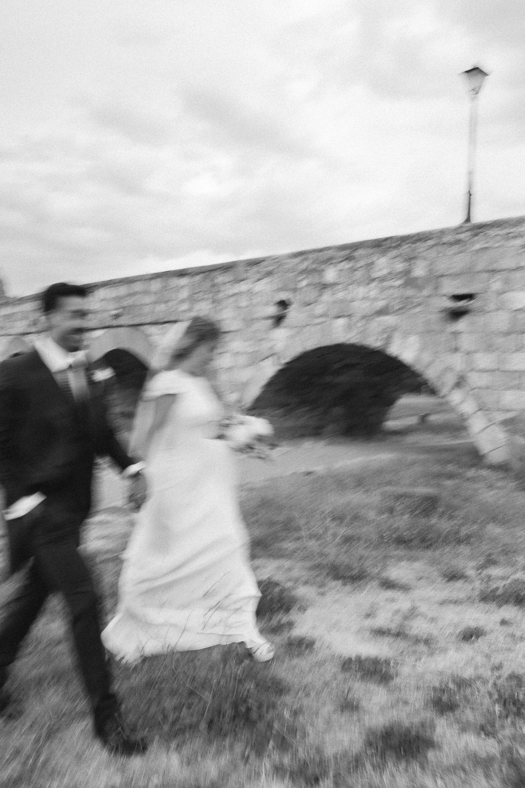 Black and white photo of a bride and groom walking hand in hand past a stone arch bridge on their wedding day.