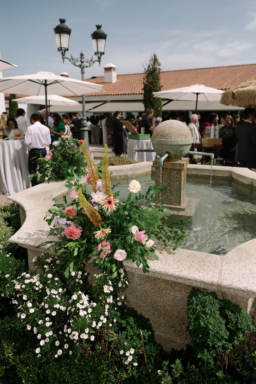 Elegant outdoor wedding reception with floral fountain centerpiece, white umbrellas, and guests mingling in a courtyard.