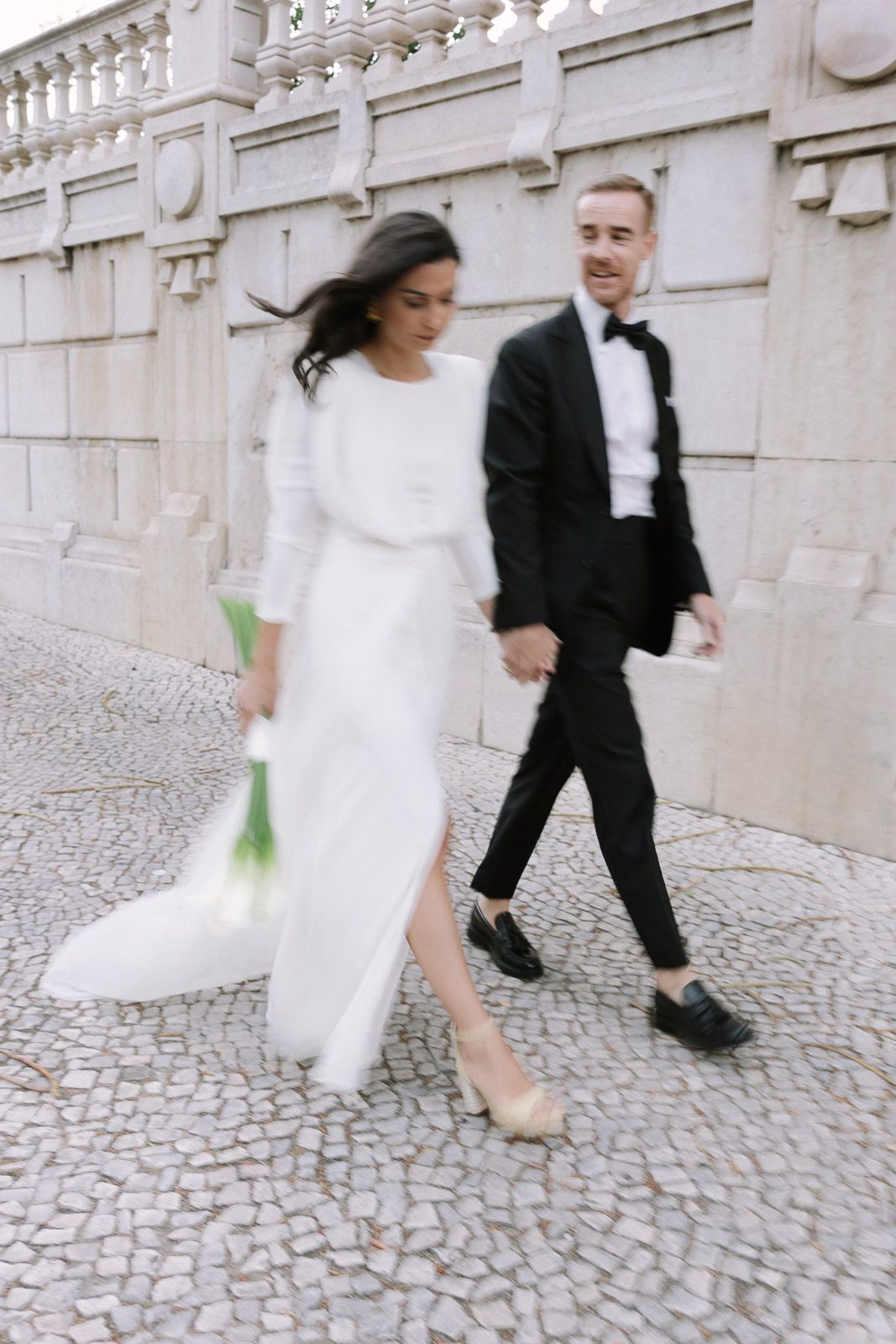 Bride and groom walking on cobblestone street, bride in white gown holding calla lilies, groom in black tuxedo.