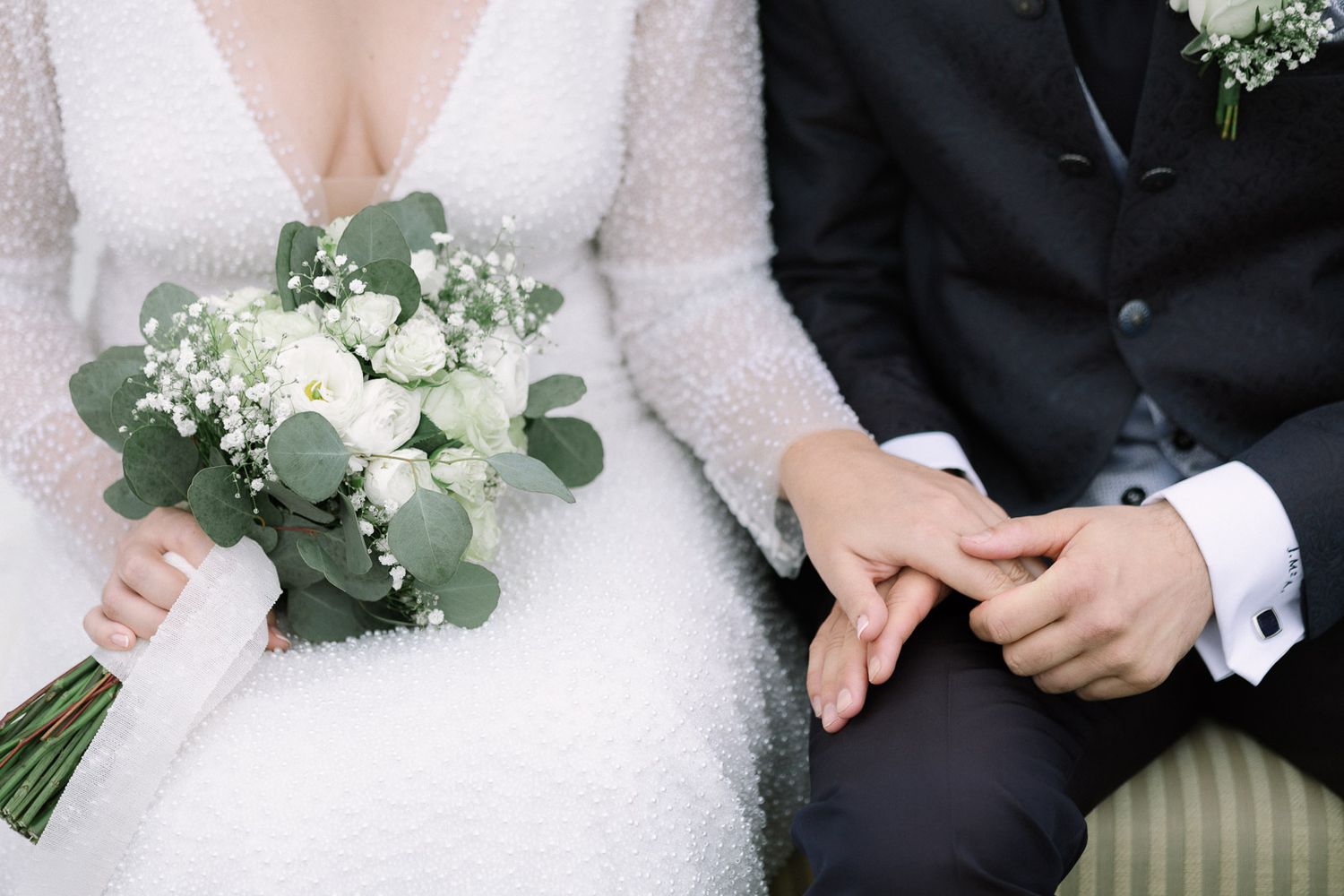 Bride holding white rose and eucalyptus bouquet while groom holds her hand, both seated in wedding attire.