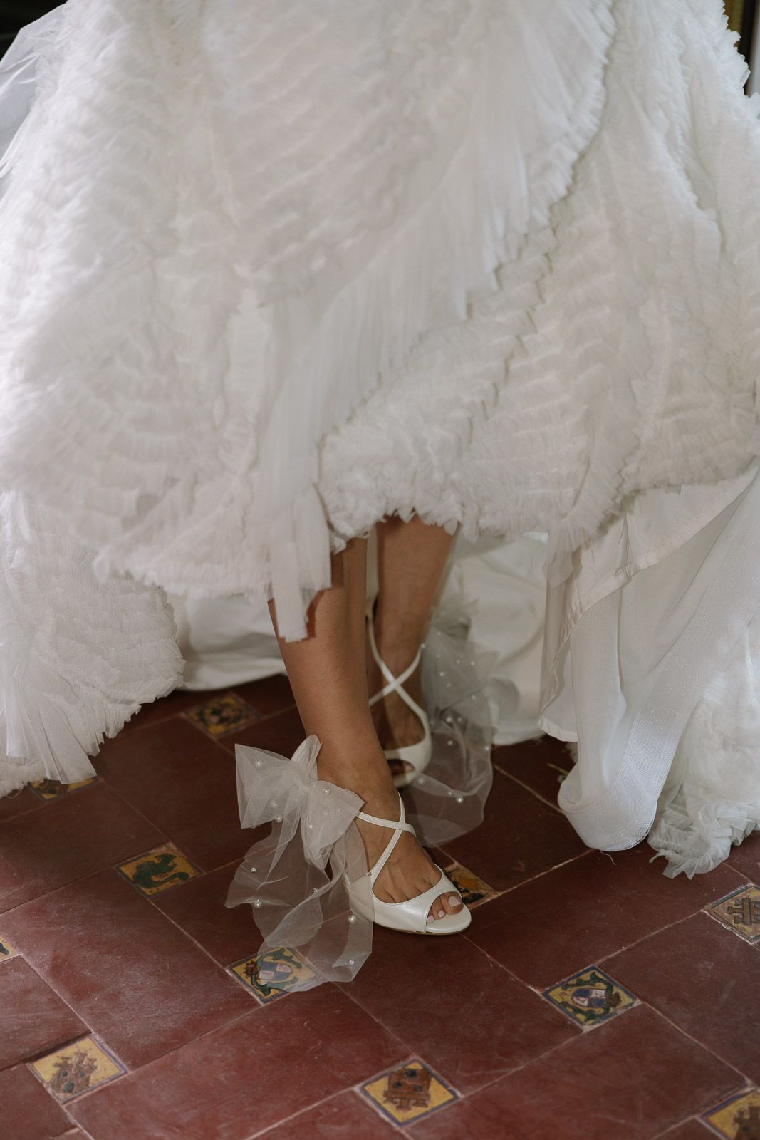 Bride adjusting elegant white wedding shoes with tulle bow details, peeking beneath a ruffled bridal gown on tiled floor.