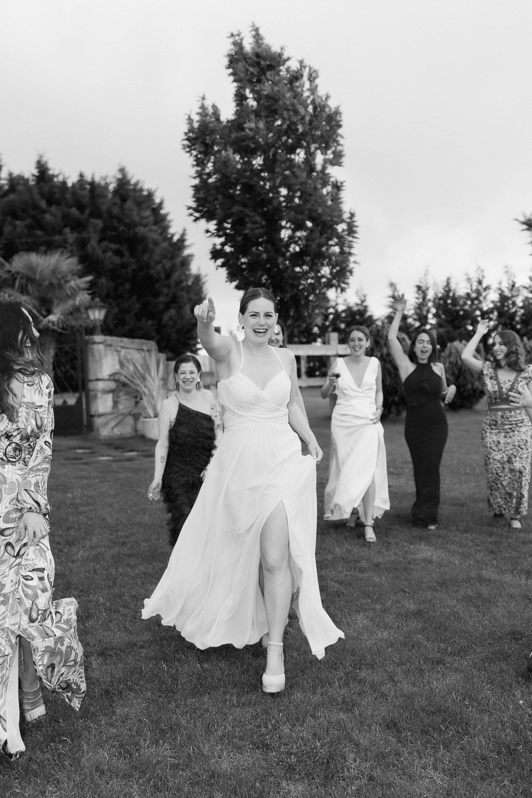 Black and white photo of a joyful bride in white gown walking with bridesmaids outdoors at a wedding celebration.