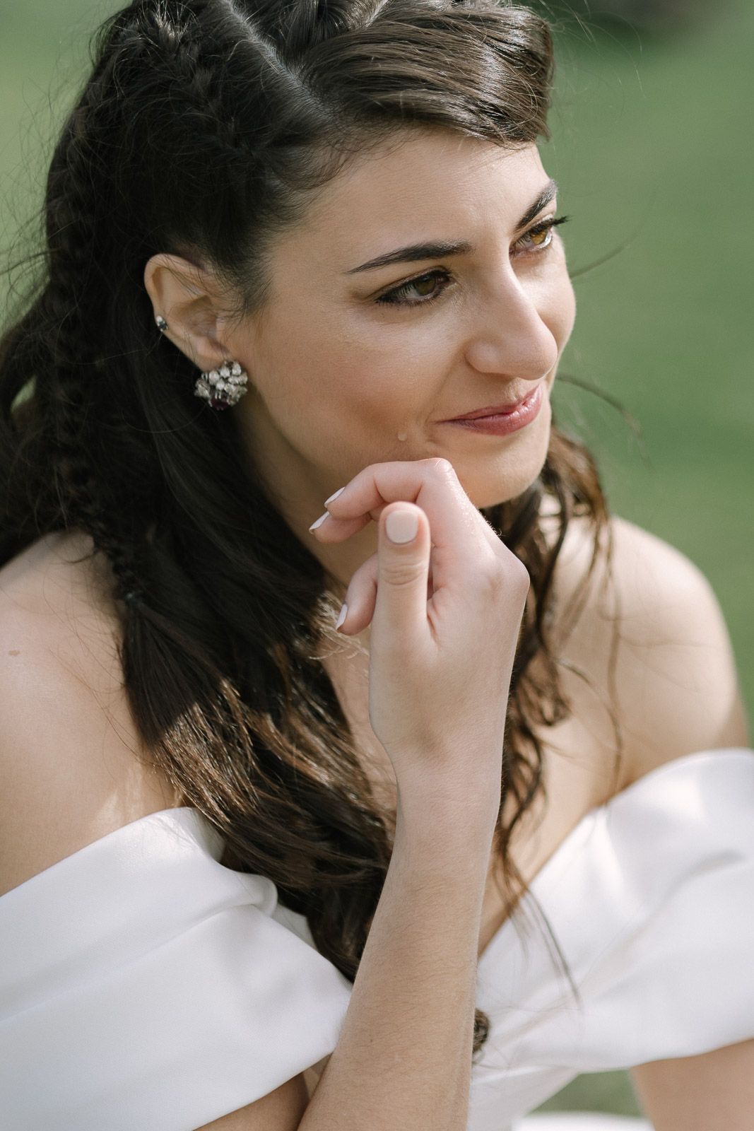 A smiling brunette bride in an off-shoulder white gown with pearl earrings, resting her chin on her hand outdoors.
