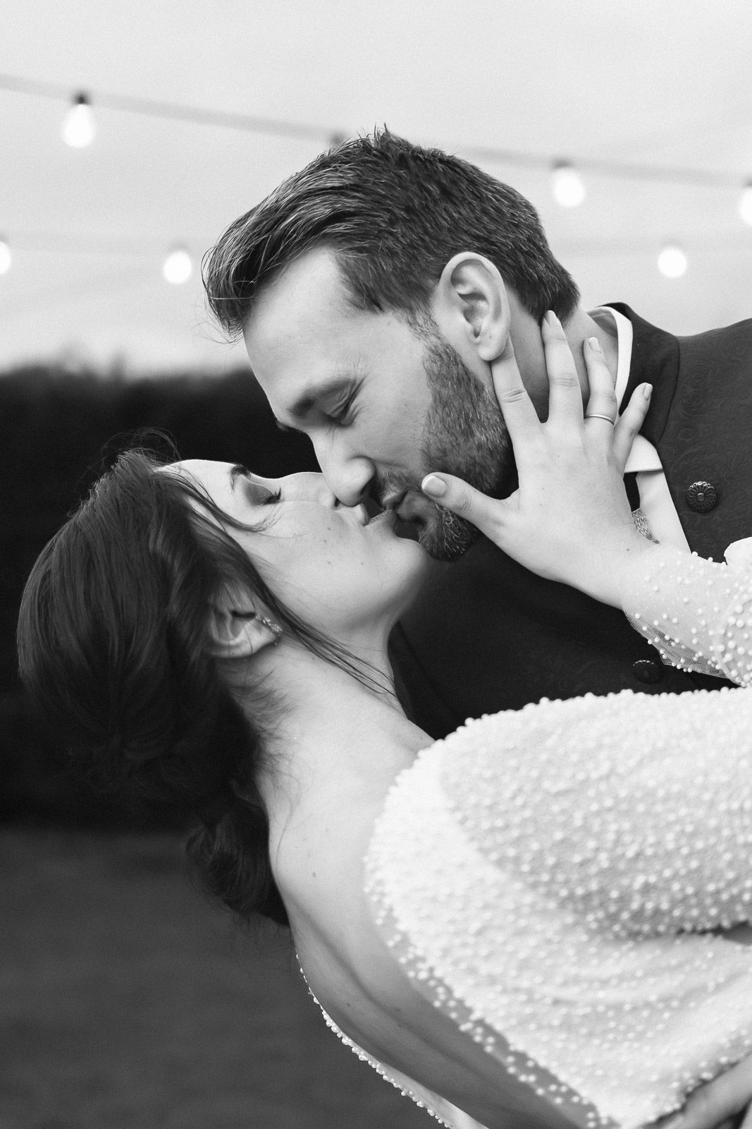 Black and white photo of a couple sharing a romantic kiss at their wedding reception under string lights.